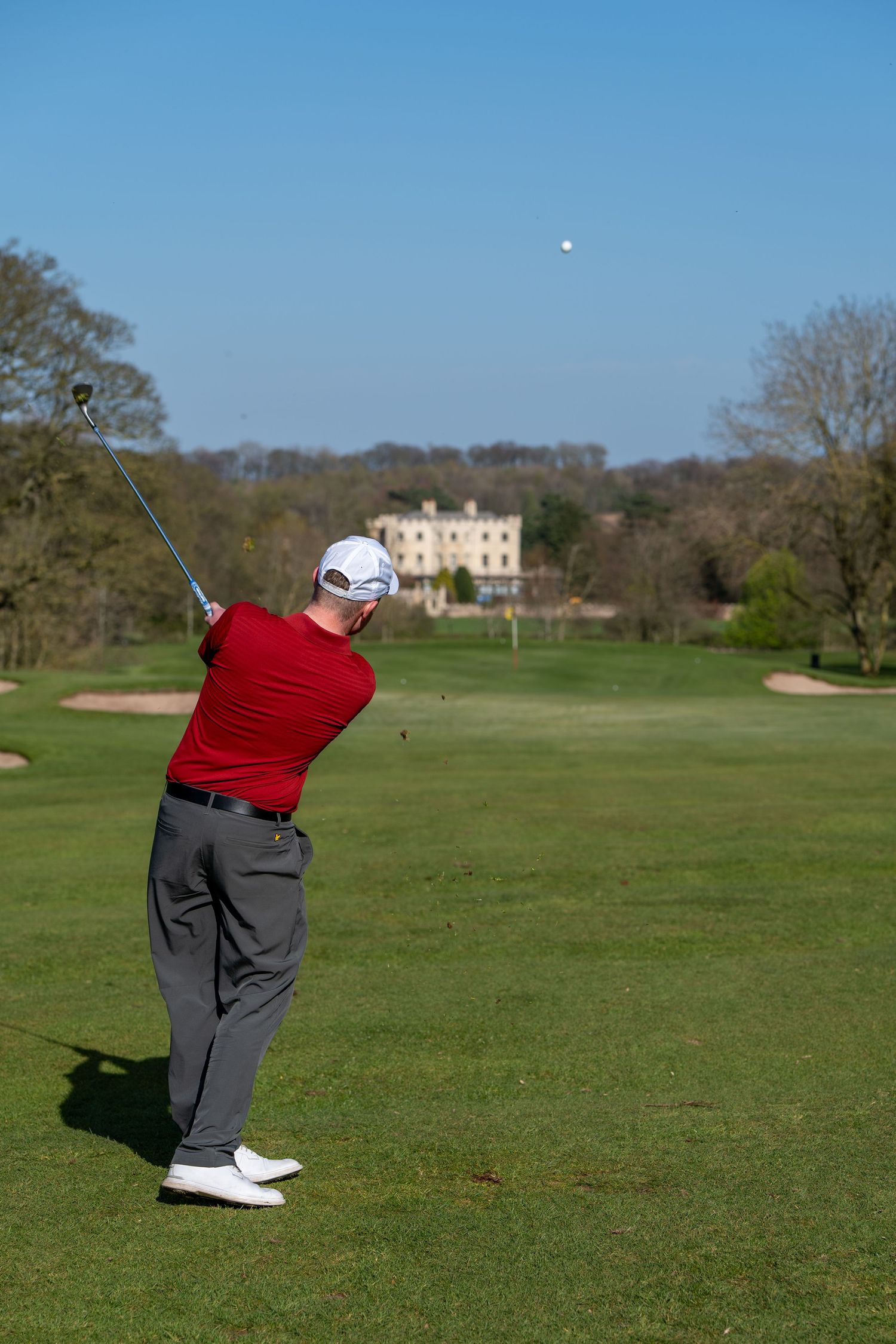 Golfer in red shirt follows through on a swing on a sunny golf course with a grand house in the background.