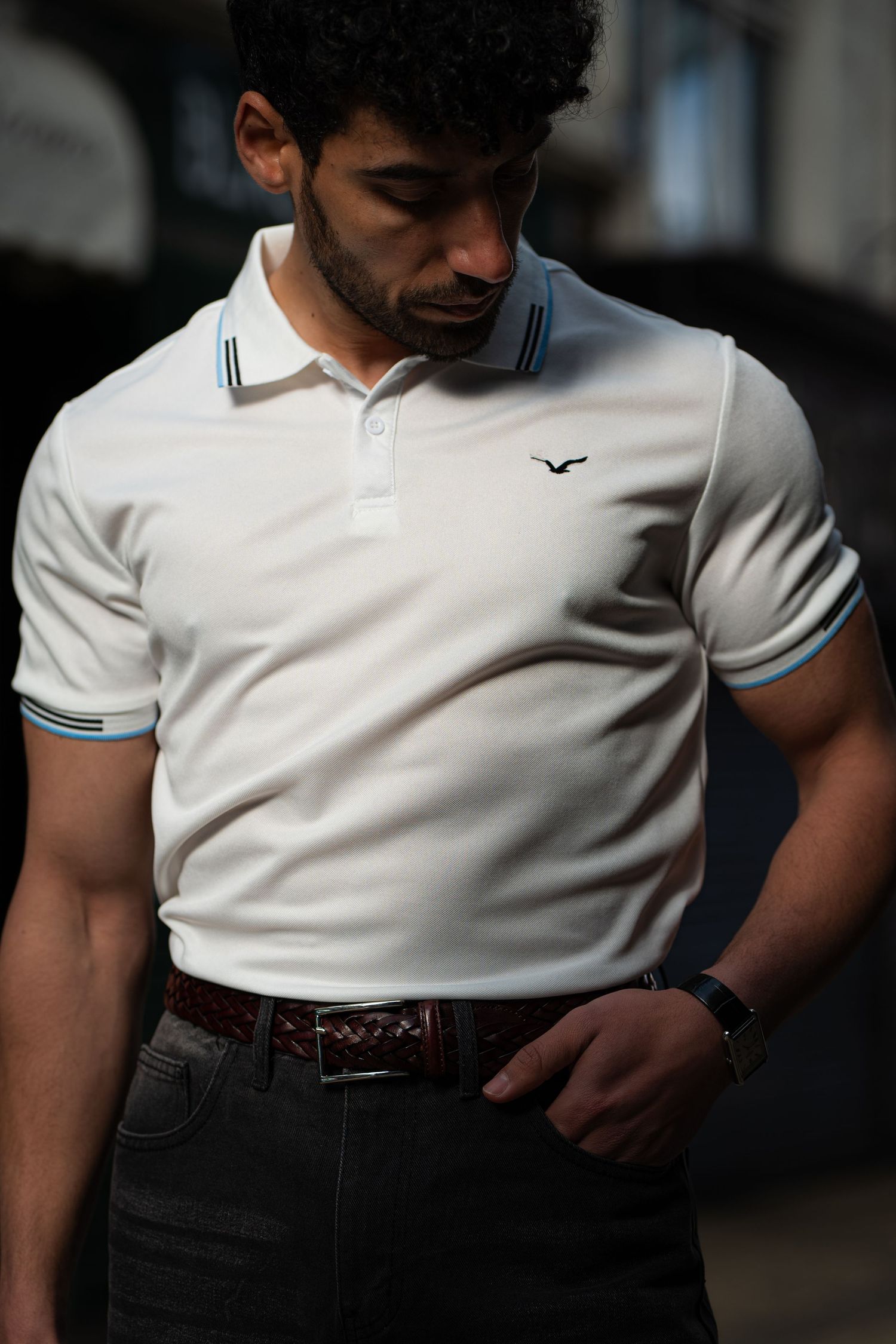 Man wearing white polo shirt with blue trim and bird logo, dark jeans, and brown belt outdoors.