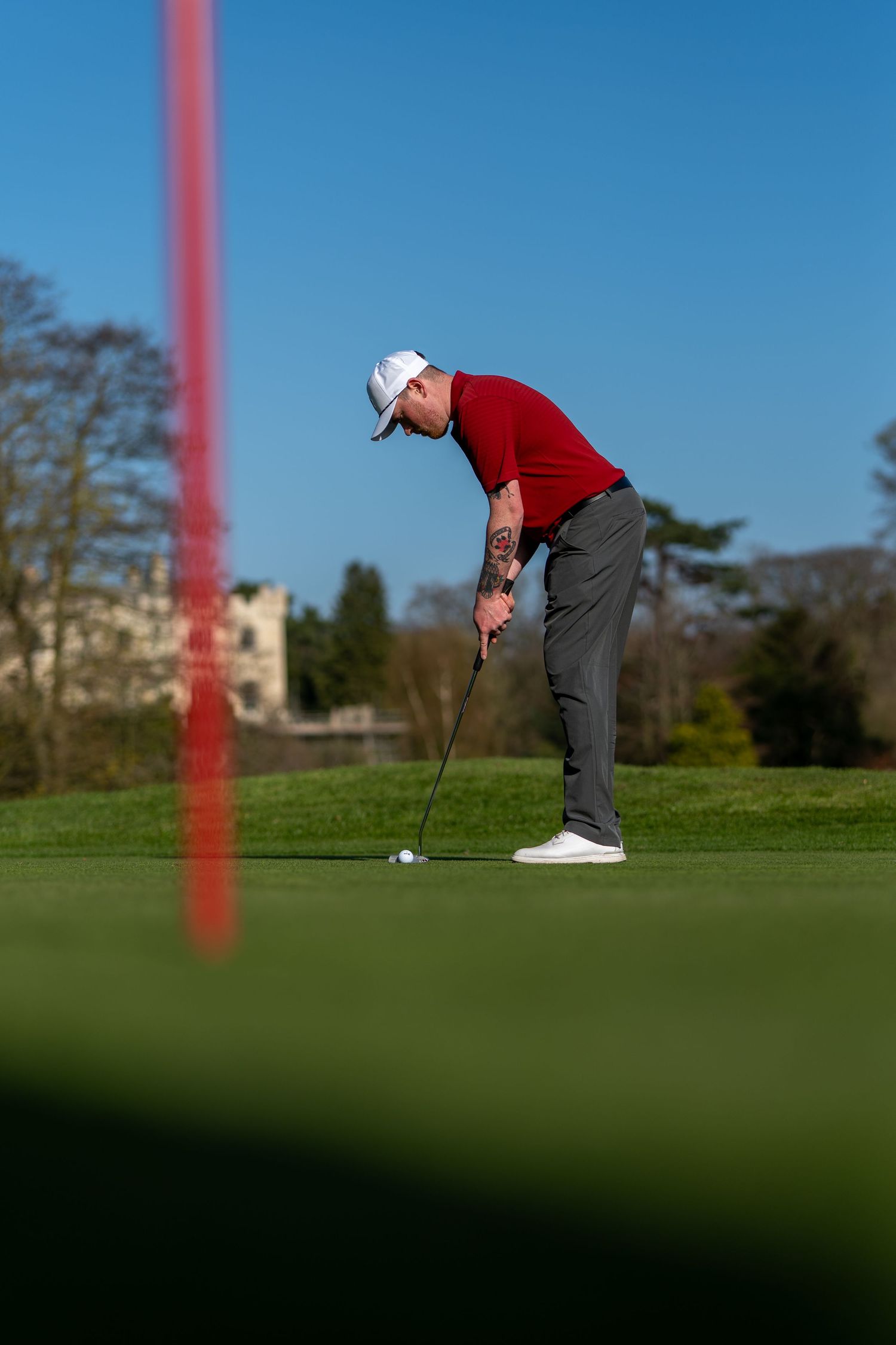 Tattooed golfer in red shirt and white cap putting on a green golf course on a sunny day.
