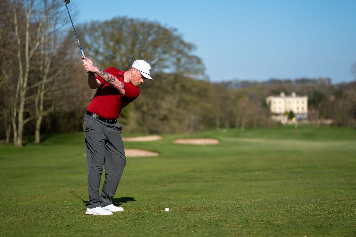 Golfer in red shirt and white cap swinging a club on a lush green golf course with trees and a building.