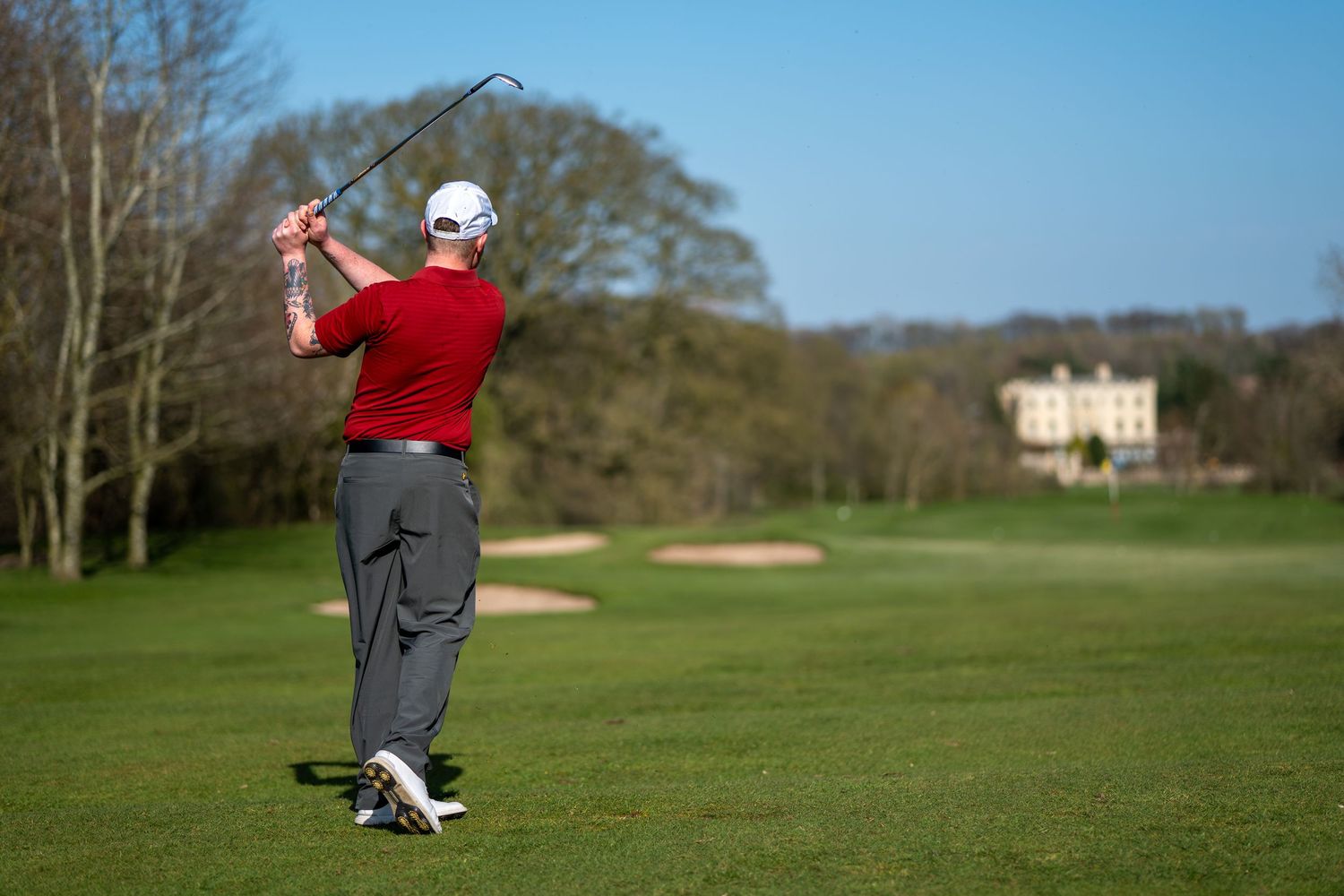 Golfer in red shirt completing a follow-through swing on a sunny golf course with a manor house in the background.