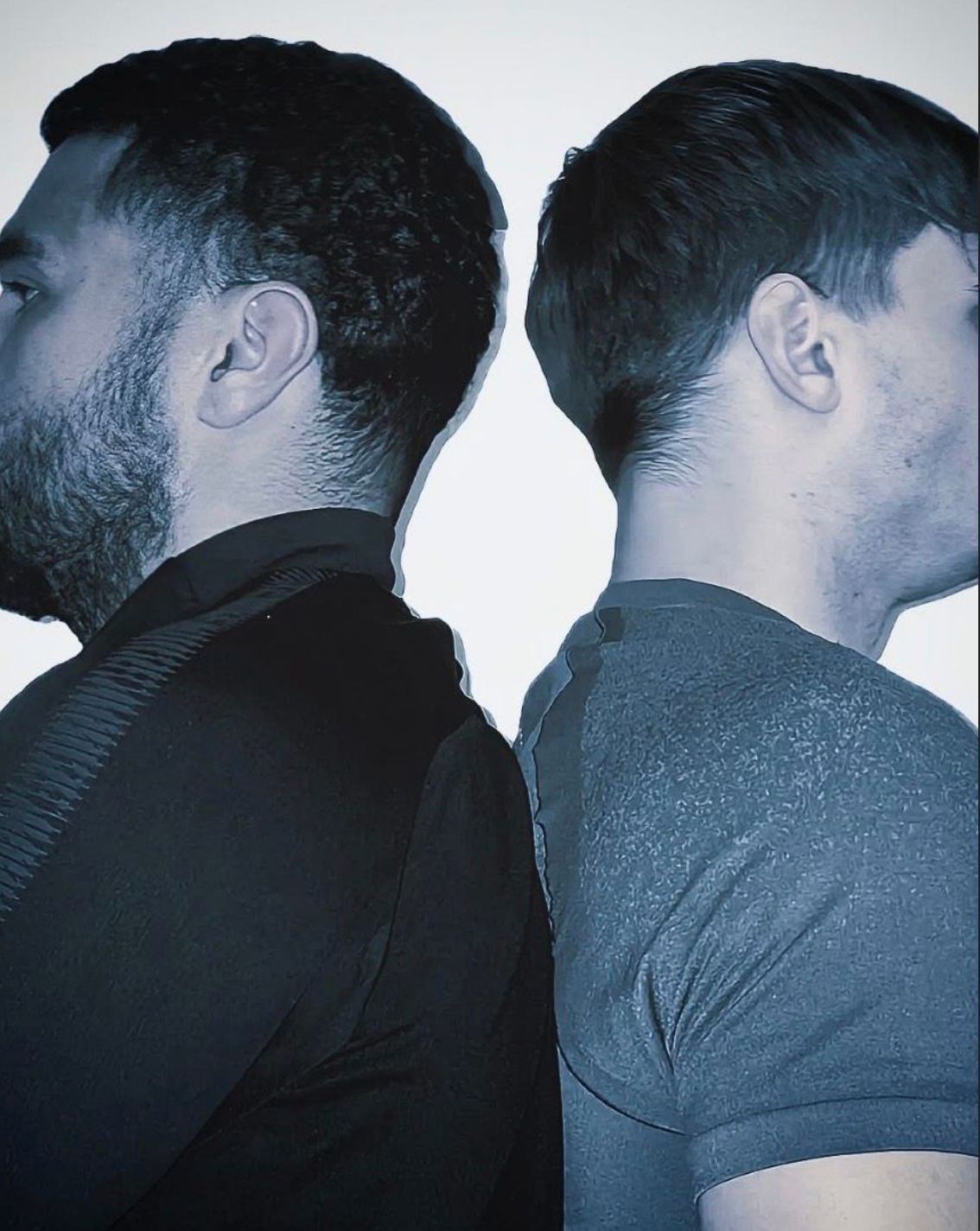 Two people stand back-to-back in an artistic portrait with one wearing black and one wearing grey.