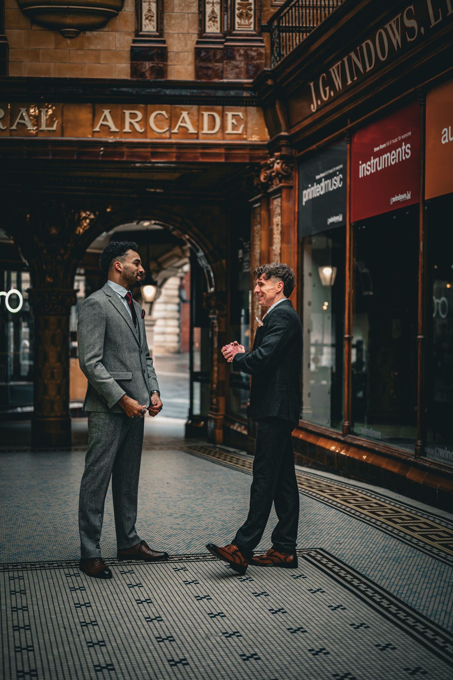 Two people in suits stand and converse in front of a vintage arcade storefront with tiled floors.