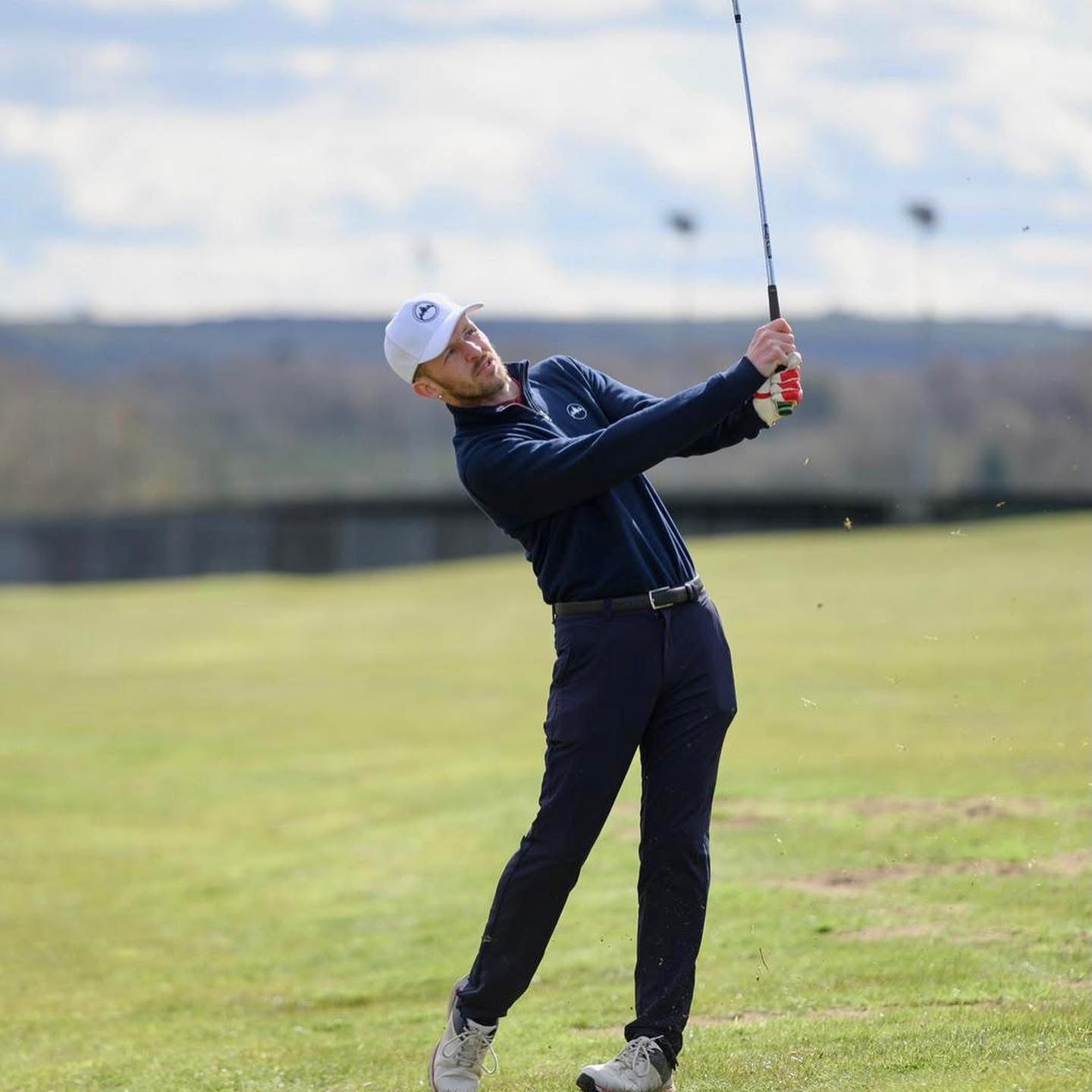 Golfer in navy outfit swings club on green course with rolling hills in background during daytime.