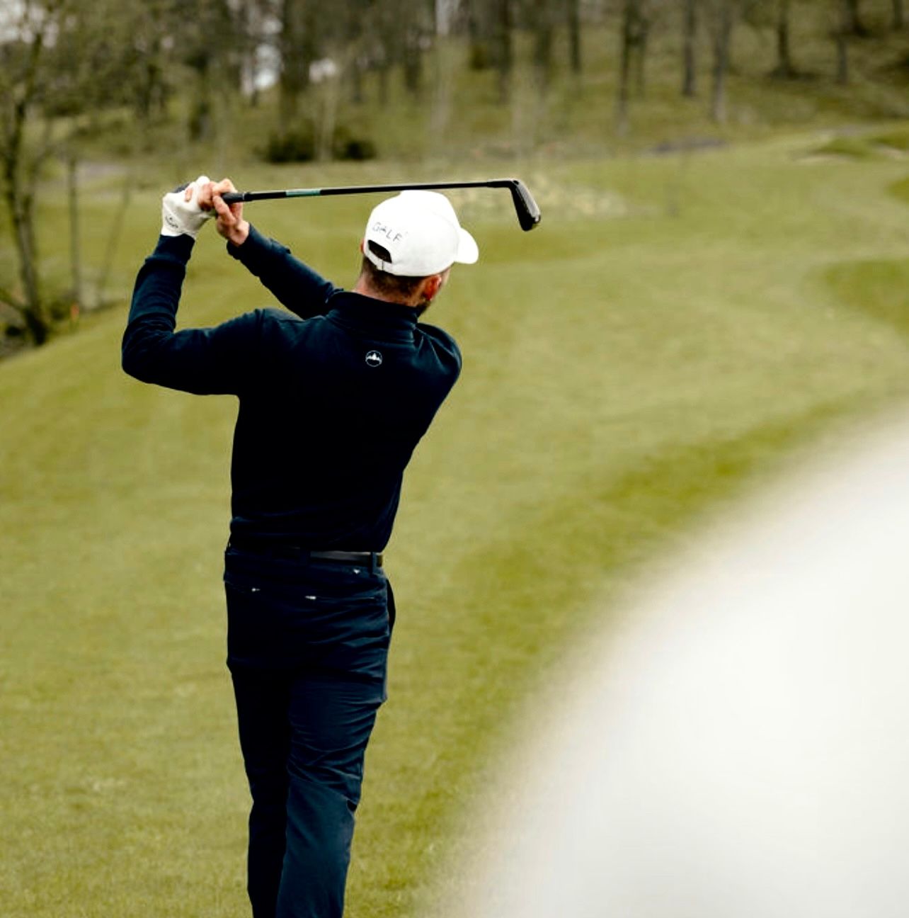 Golfer in navy sweater and white cap takes backswing on scenic course with trees in background.