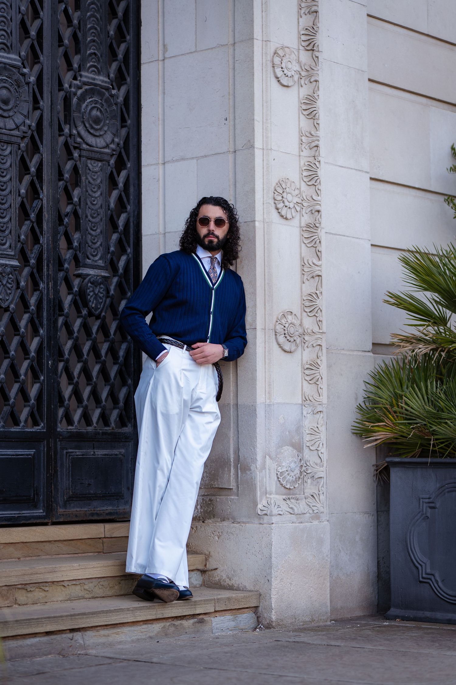 Man in navy cardigan and white wide-leg trousers leaning against ornate stone building entrance with decorative door.