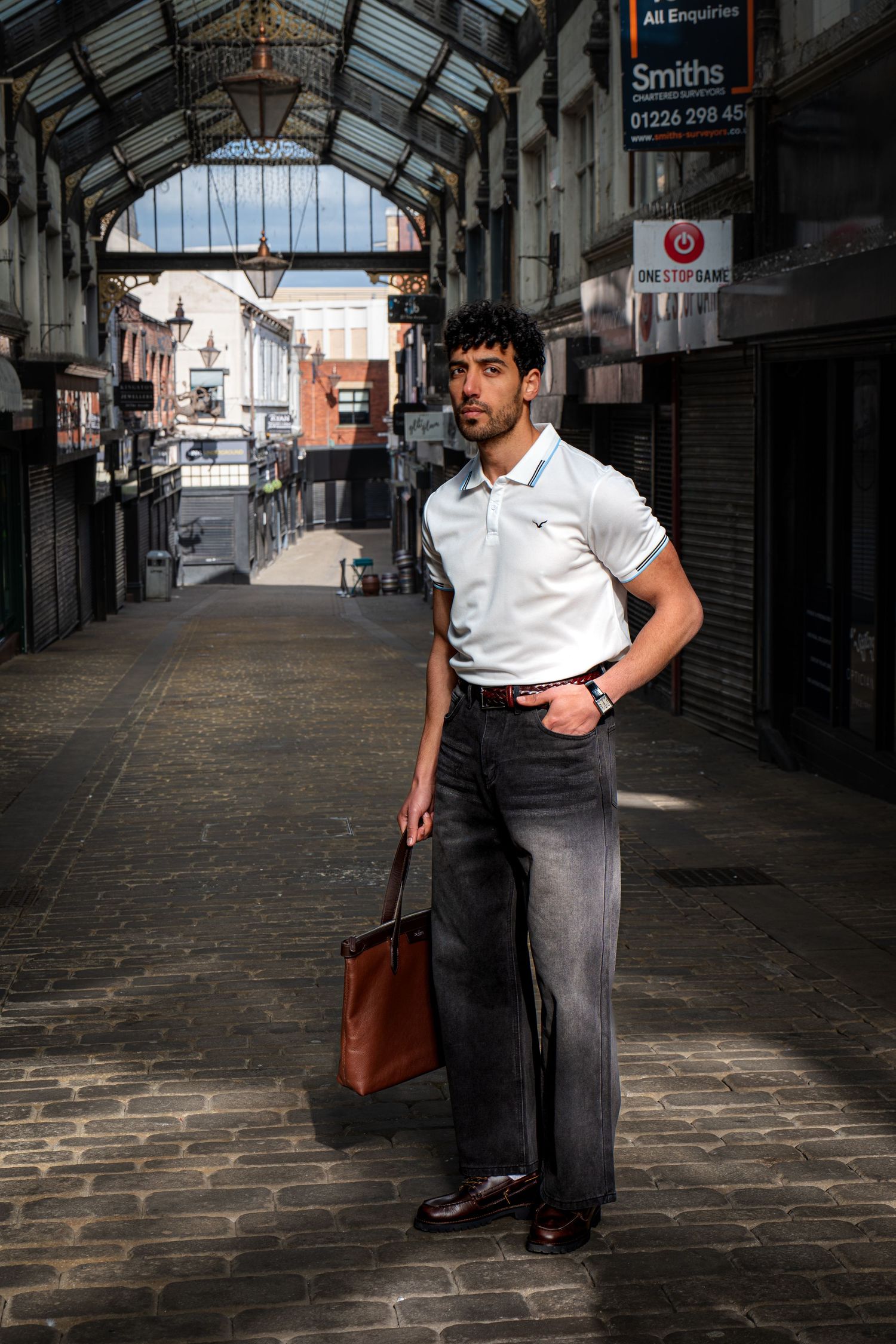 Stylish man in white polo and dark wide-leg jeans carrying brown leather bag in covered market.