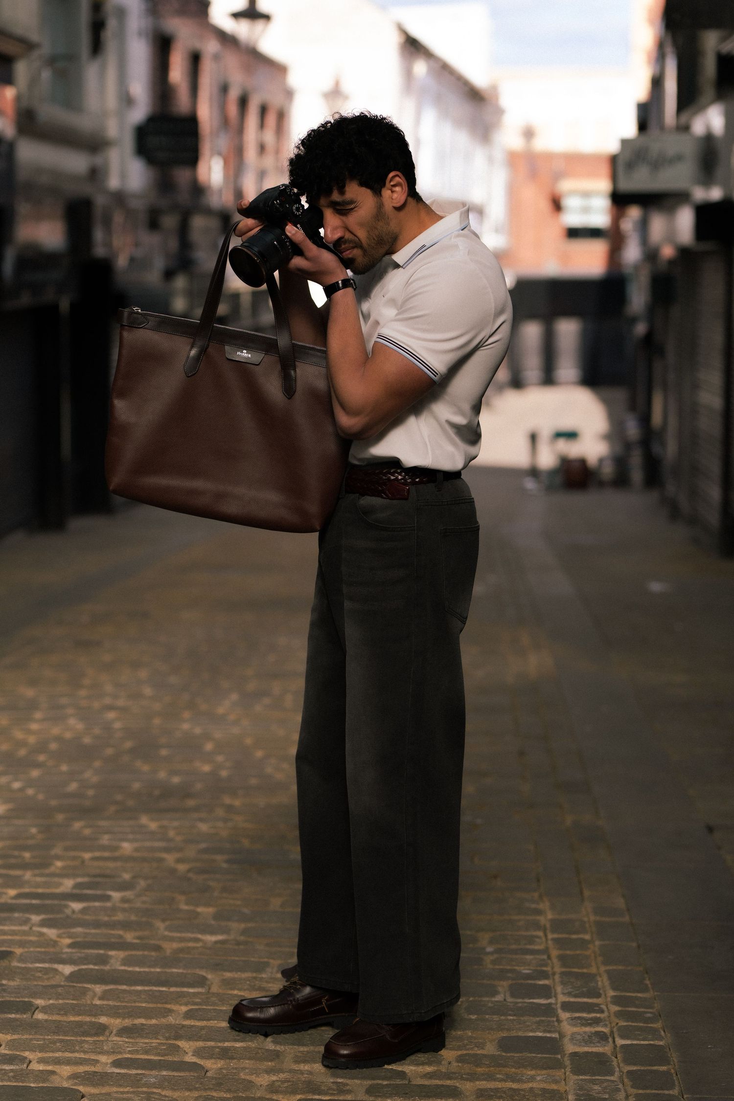 Man in white polo shirt photographing with camera while holding brown leather tote bag on urban cobblestone alley.