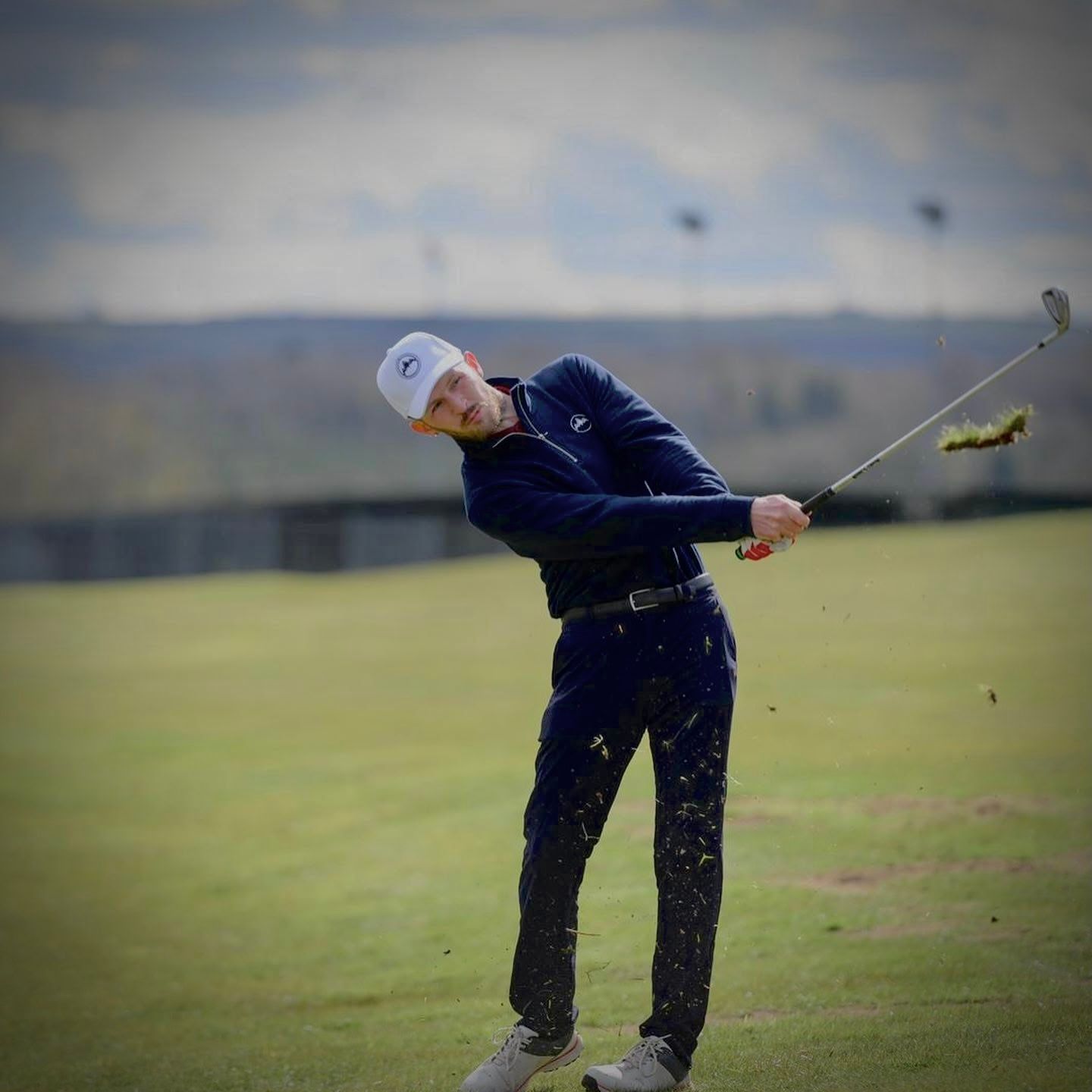 A golfer follows through on their swing while practicing on a grassy course under cloudy skies.