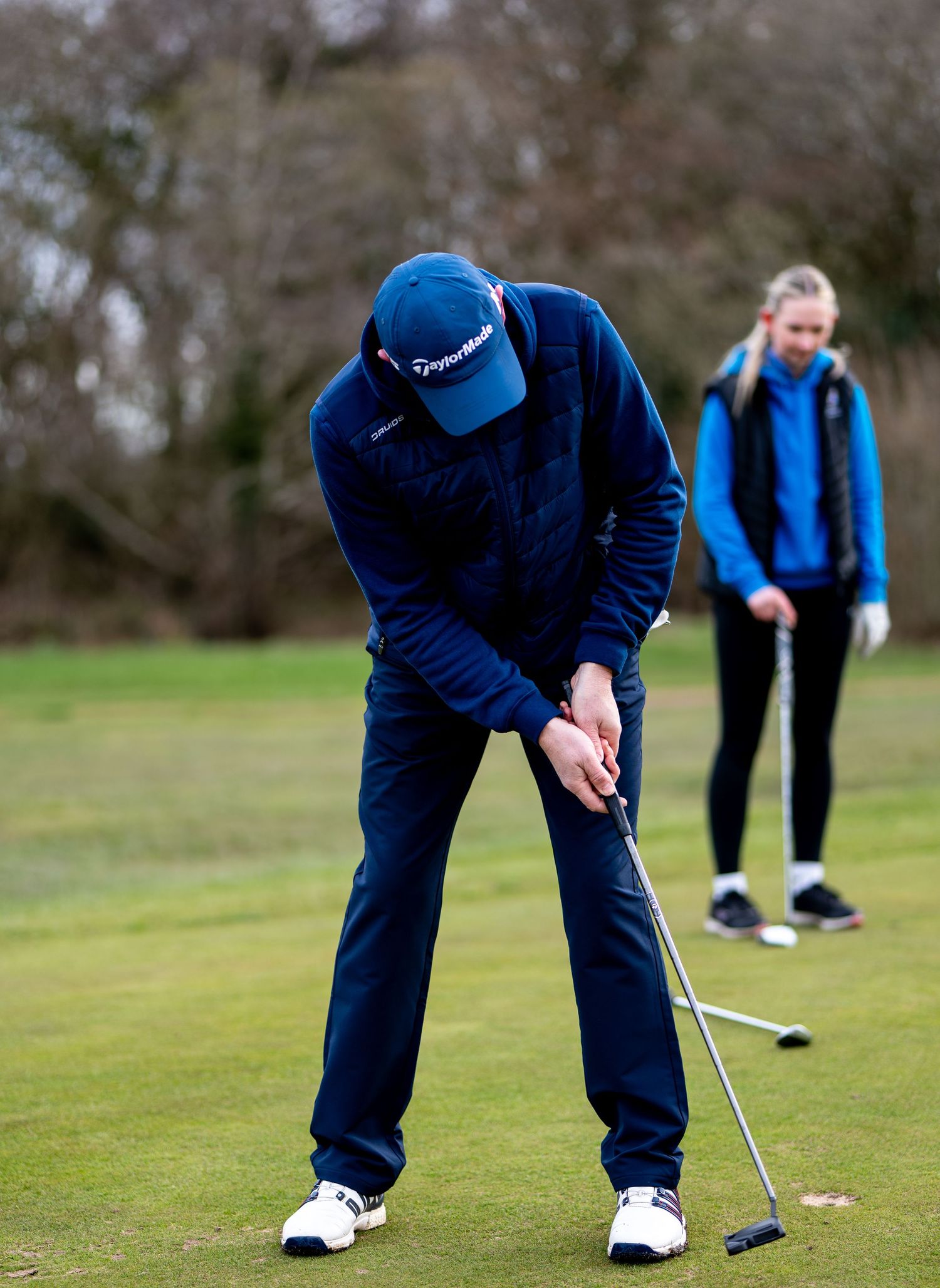 Golfer in blue jacket and TaylorMade cap putting on green while female golfer watches in background.