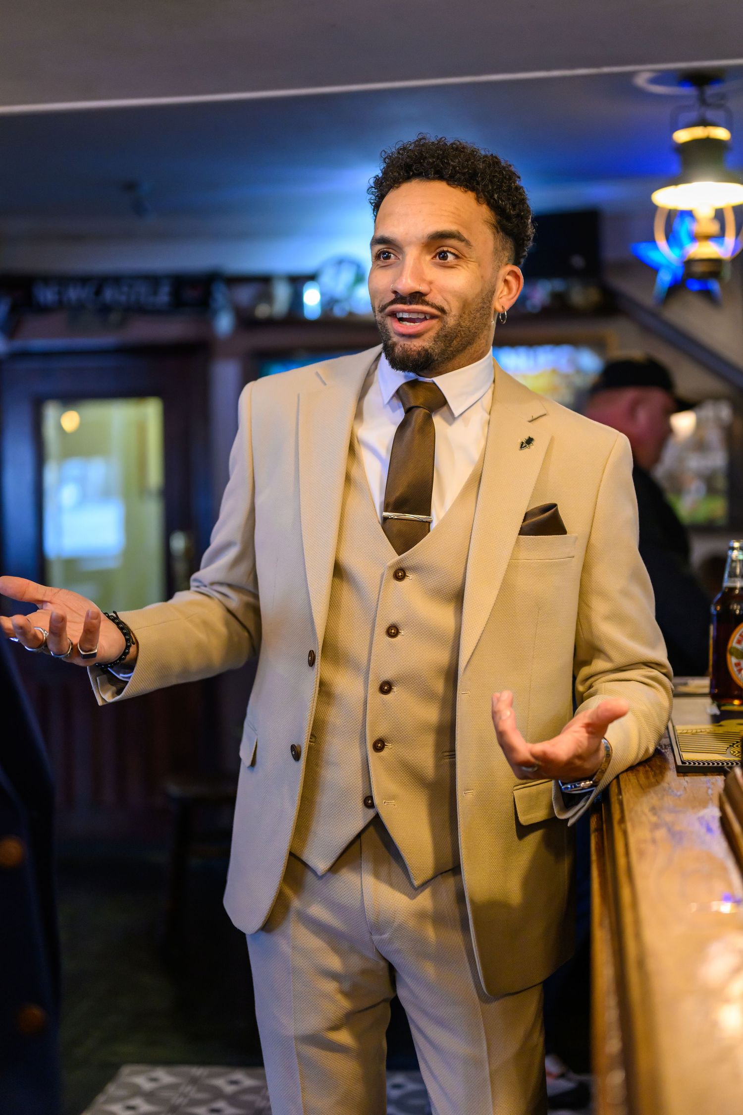 A person in a beige three-piece suit gestures animatedly while speaking in what appears to be a restaurant or bar setting.