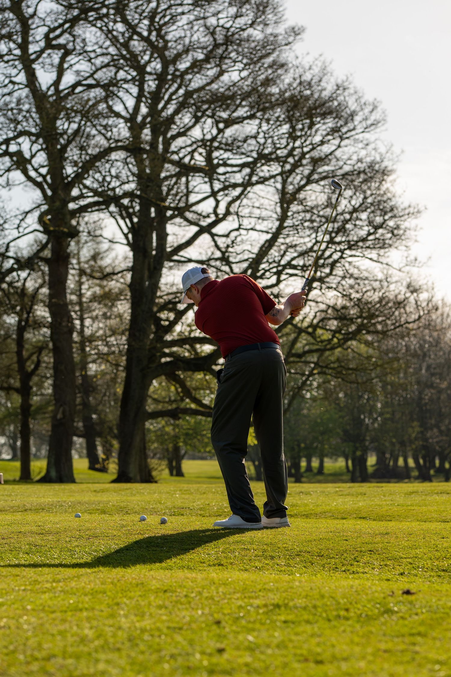 Golfer in red sweater completing a swing on a lush green golf course with bare trees in the background.