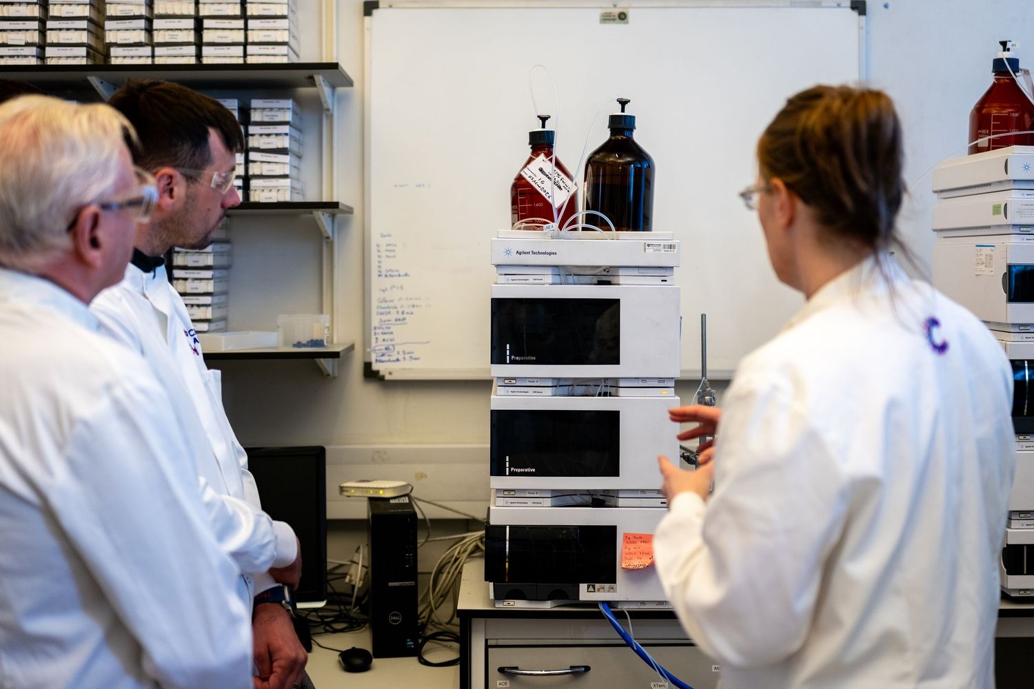Scientists in white lab coats examine laboratory equipment including an HPLC machine with reagent bottles in a research lab.