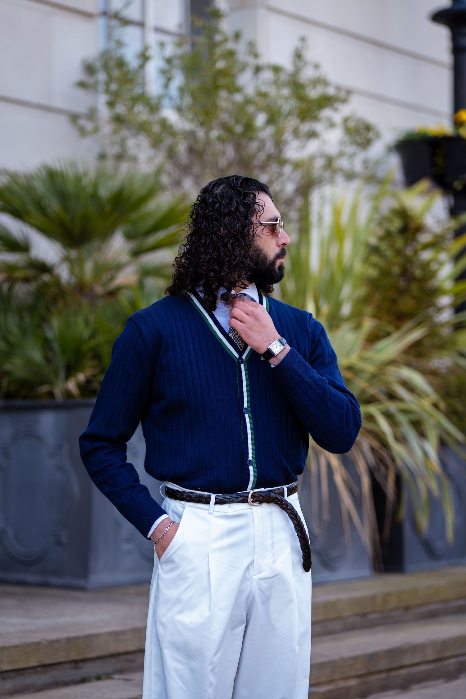 Stylish man in navy ribbed cardigan and white trousers adjusting tie outdoors with tropical plants behind him.
