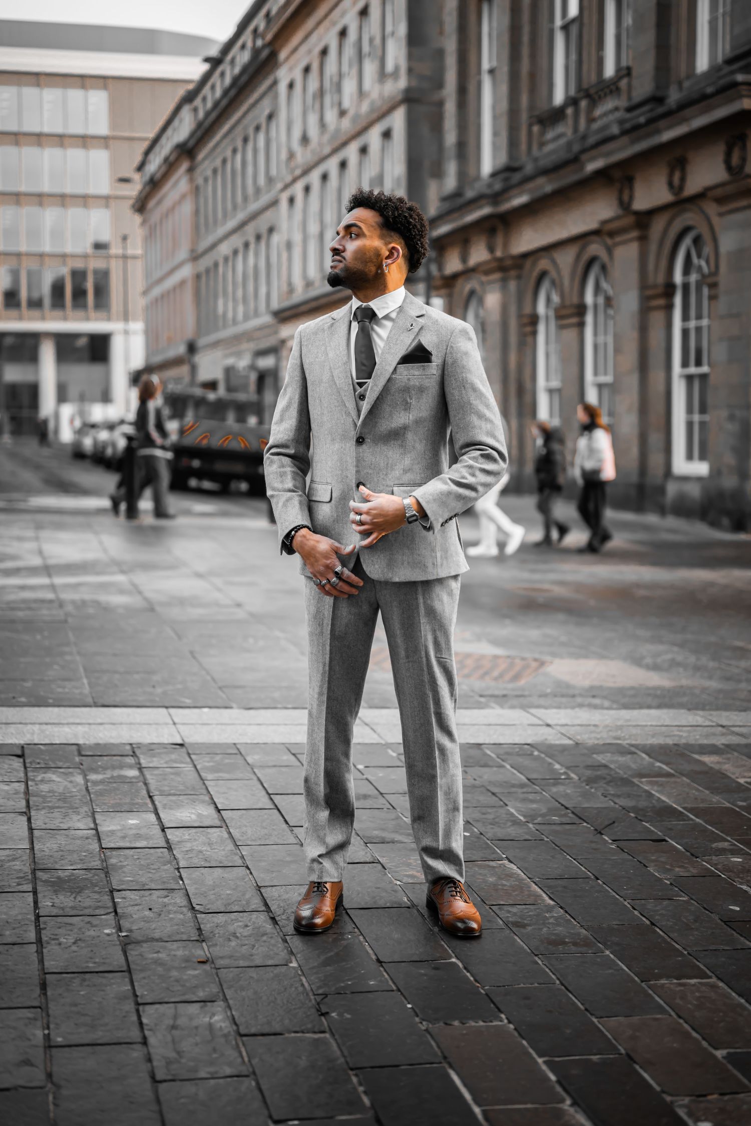 A well-dressed person in a light gray suit and brown shoes poses on a cobblestone street near historic buildings.