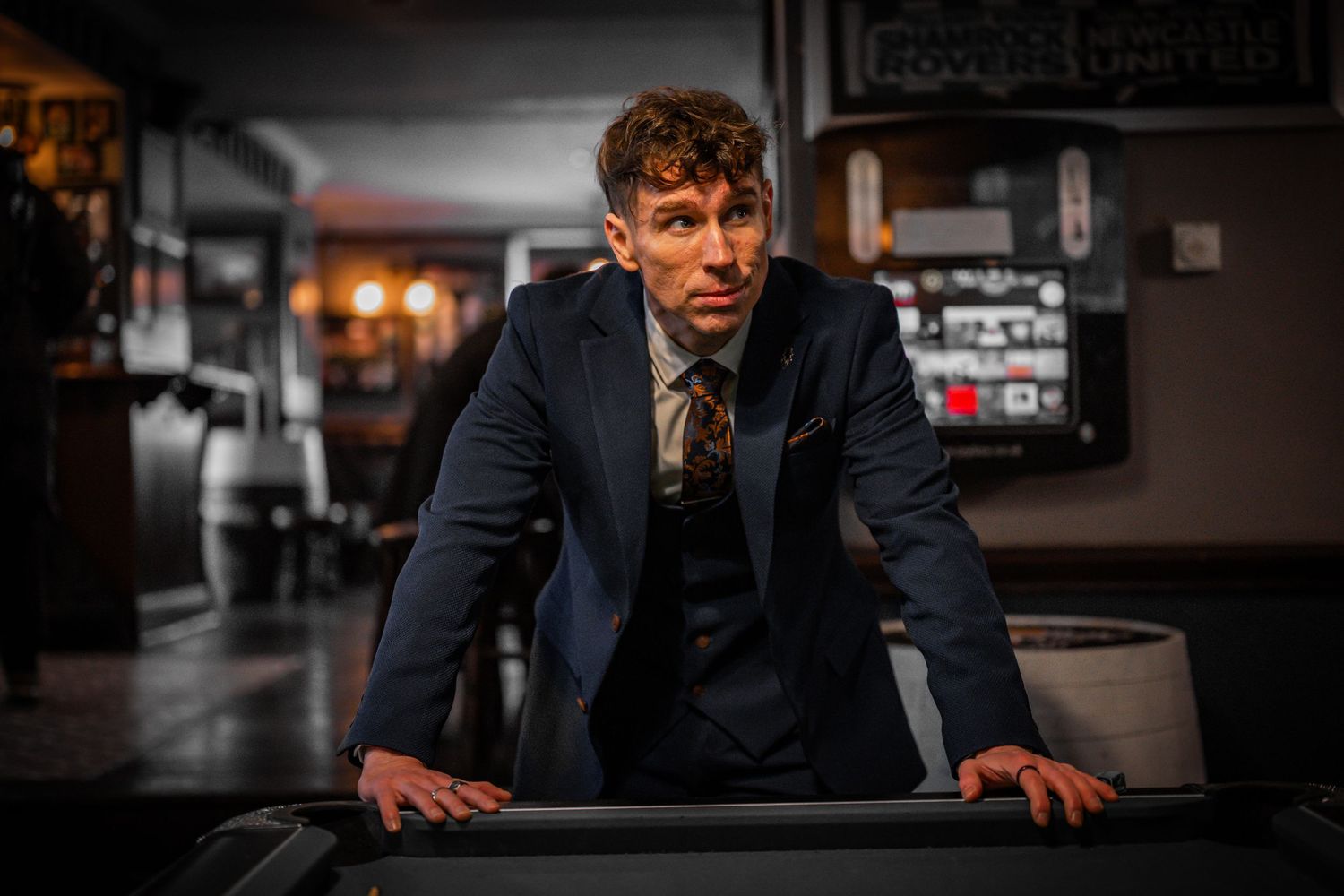 A person in a dark suit leans over a dimly lit bar counter in a moody atmospheric setting.