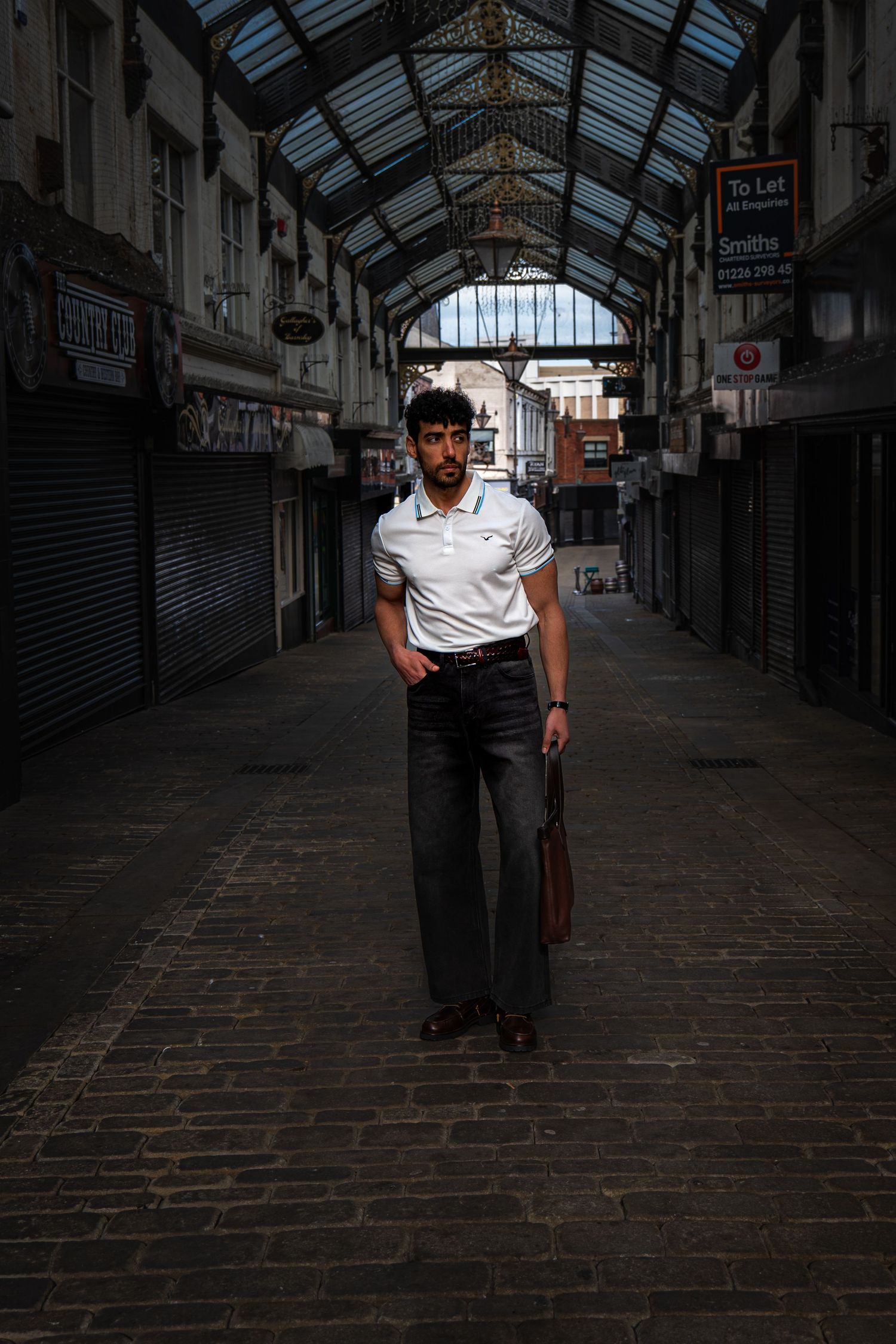 Man in white polo shirt and gray wide-leg jeans standing in a dark covered market arcade with cobblestone floor.