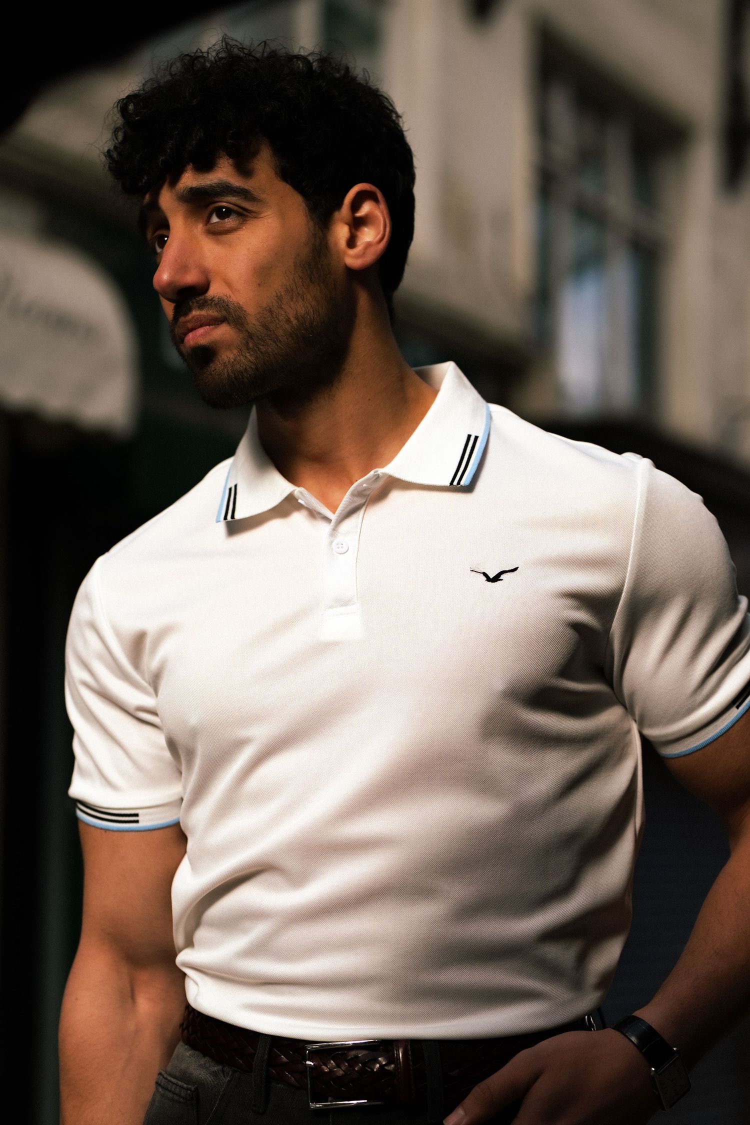 Muscular man in white Hollister polo shirt with black curly hair posing outdoors.
