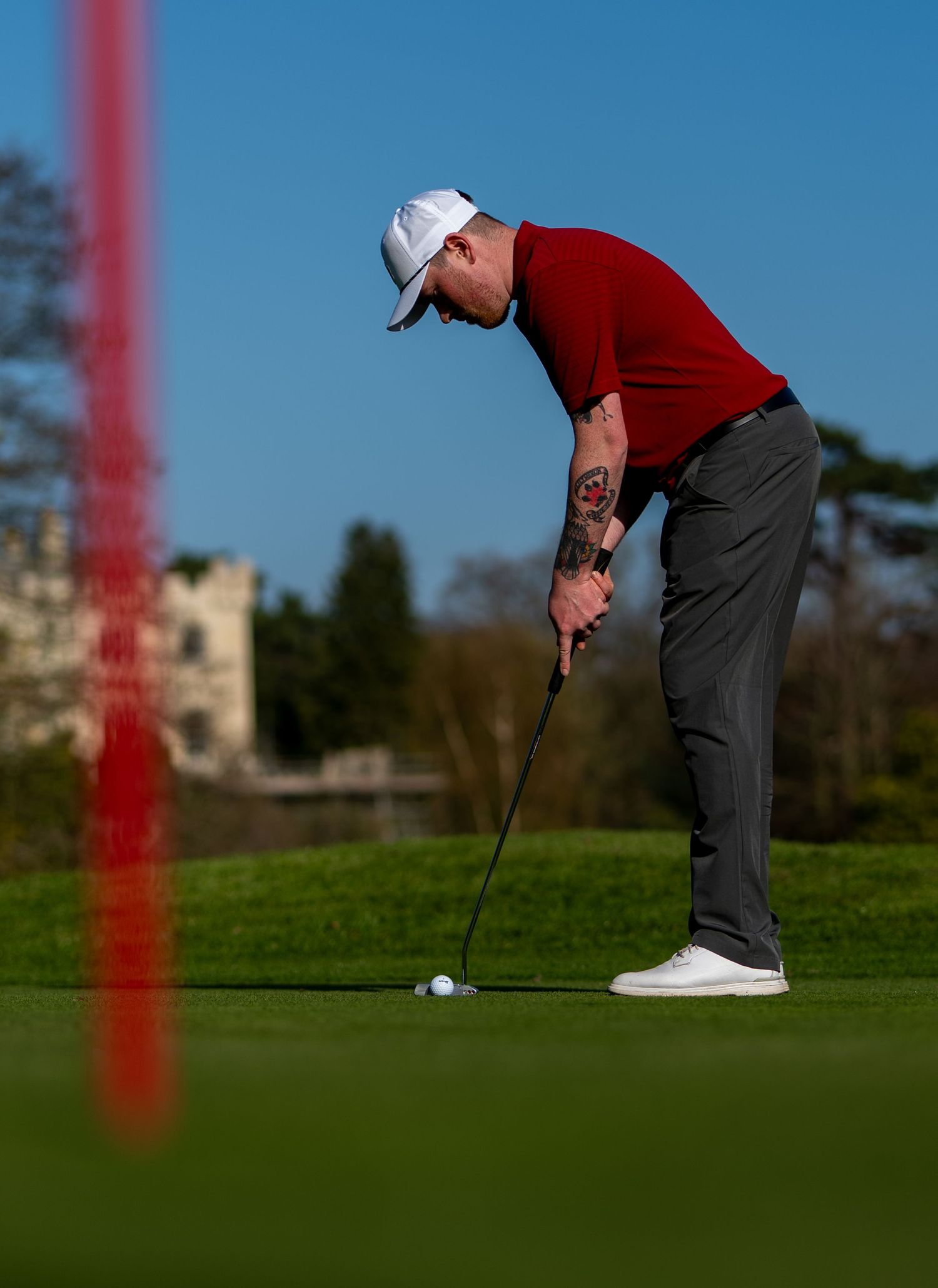 Golfer in red sweater and white cap putting on a green course with a castle in the background on a sunny day.