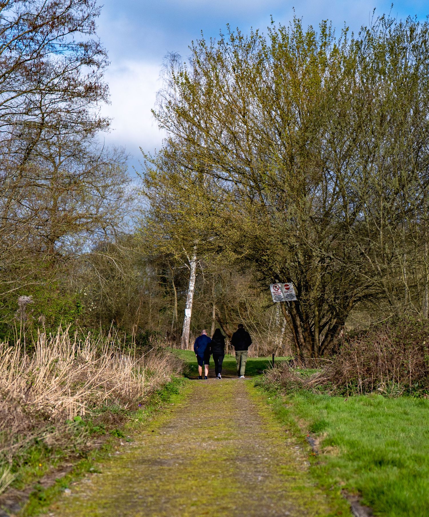 Two people walking along a narrow path through a lush green woodland nature reserve on a bright spring day.