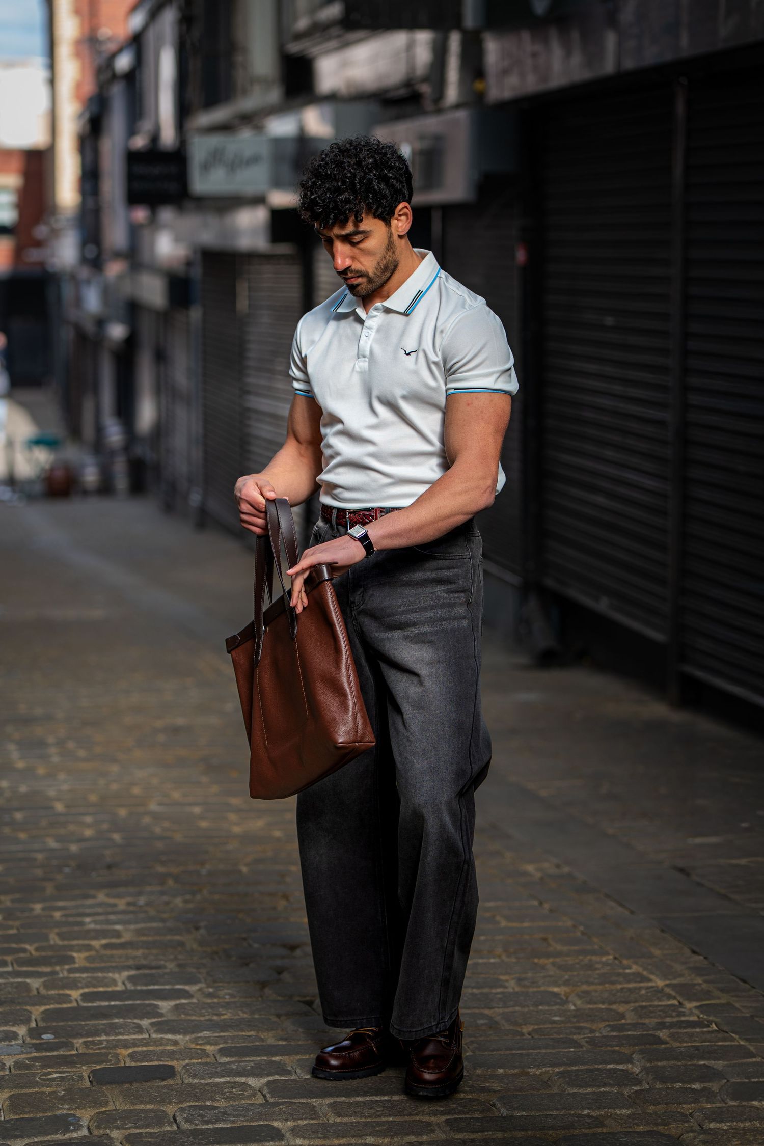 Man in white polo shirt holding brown leather tote bag on cobblestone alley street.