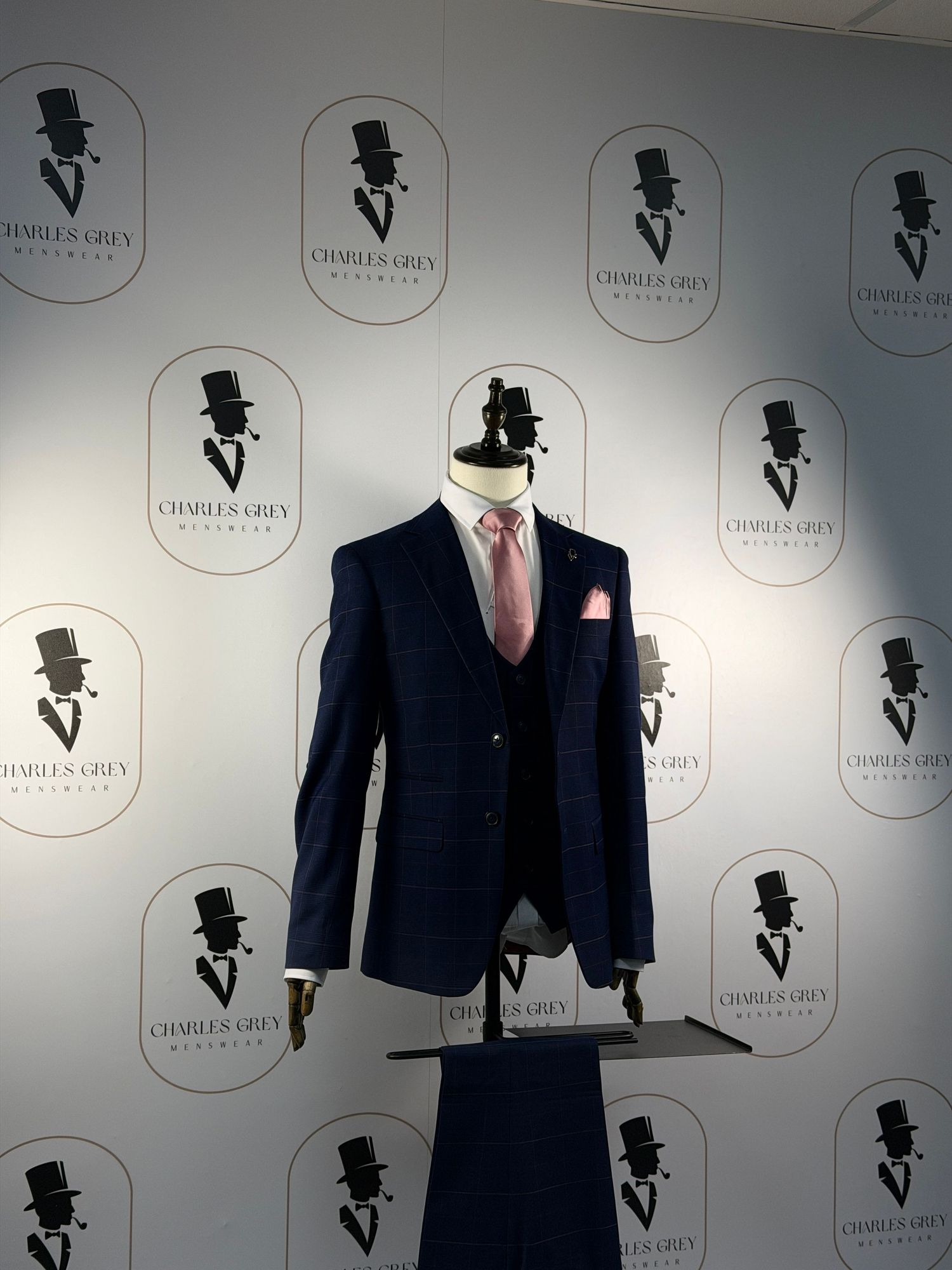 Display of formal menswear suits and jackets against a branded wall with top hat logo repeating pattern.