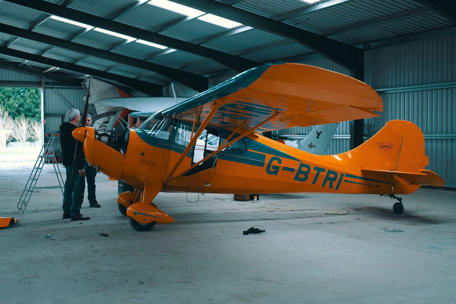 Four vintage orange Piper Cub aircraft parked in an aircraft hangar with fluorescent lighting overhead.