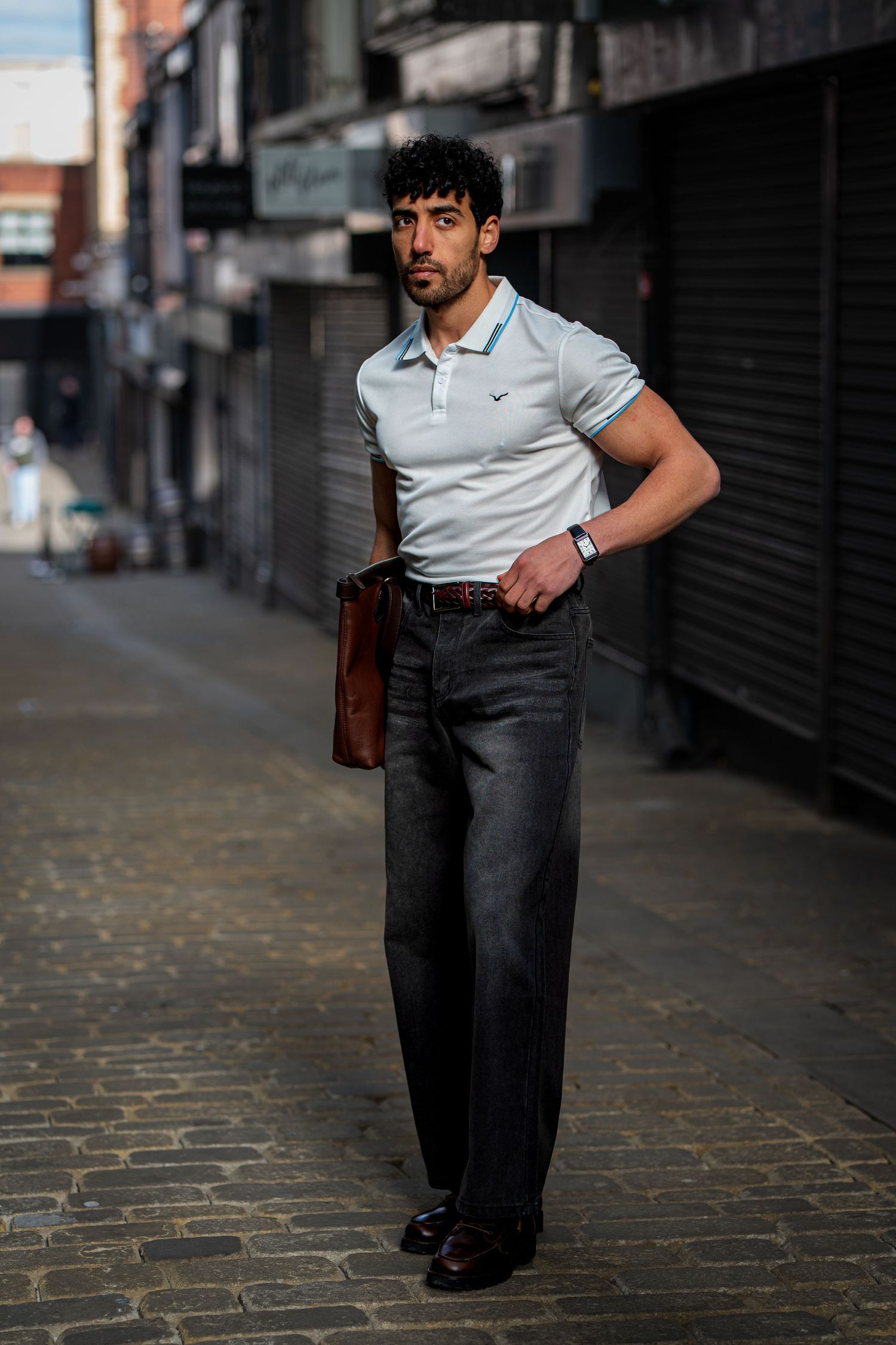 Man in white polo shirt and dark wide-leg jeans holding brown bag on cobblestone alley.