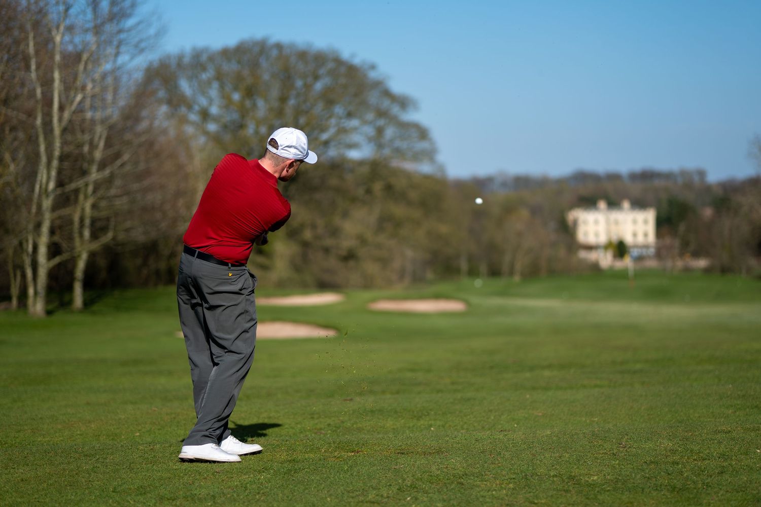 Golfer in red shirt and white cap swinging a club on a lush green golf course with a mansion in the background.