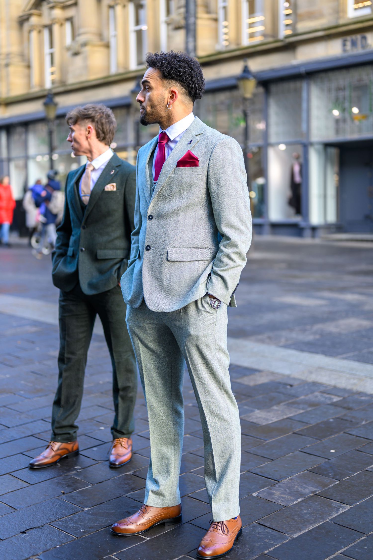 Two men in stylish suits standing on a city street, one wearing light gray with a red tie and one in dark green.