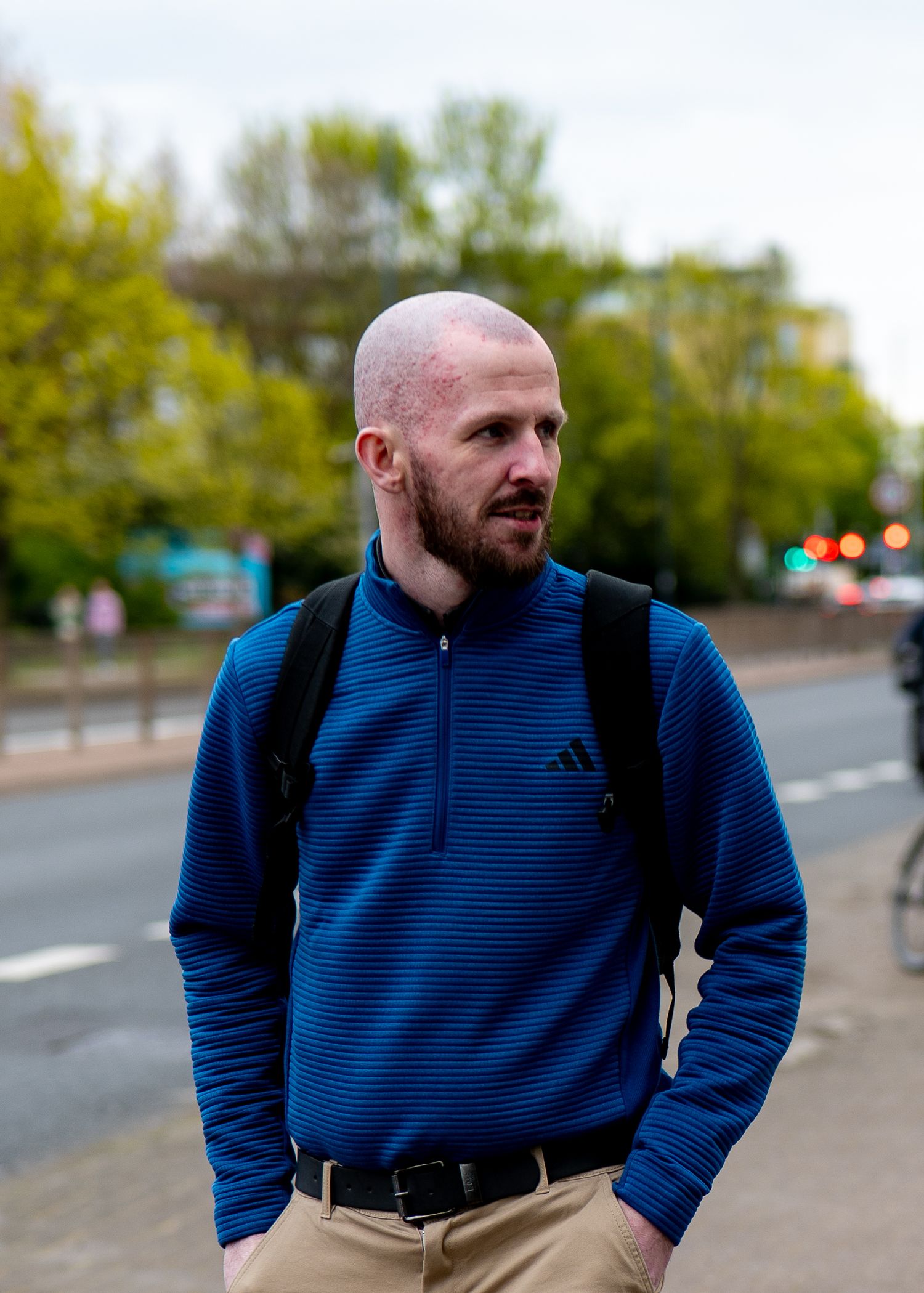 Bald man with beard wearing blue Adidas zip-up pullover and backpack standing on urban street.