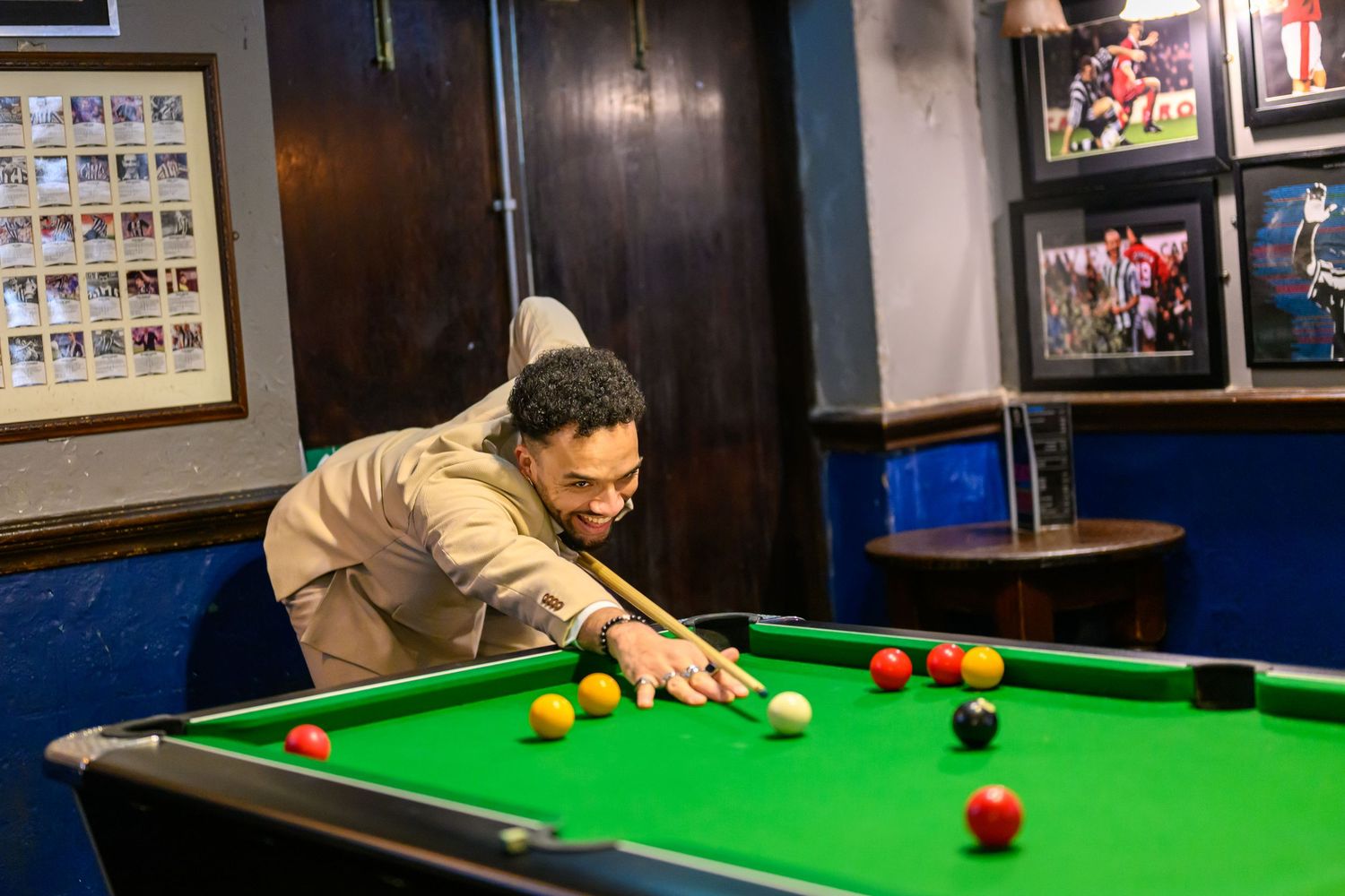 A player lines up a shot on a green pool table in a dimly lit bar with framed photos on the wall.