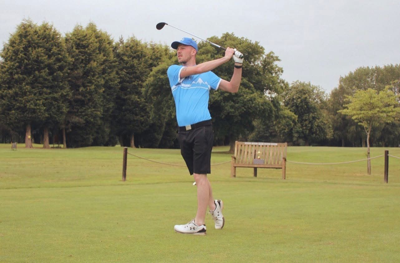 A golfer in a black polo shirt swings a club on a lush green golf course with trees in the background.