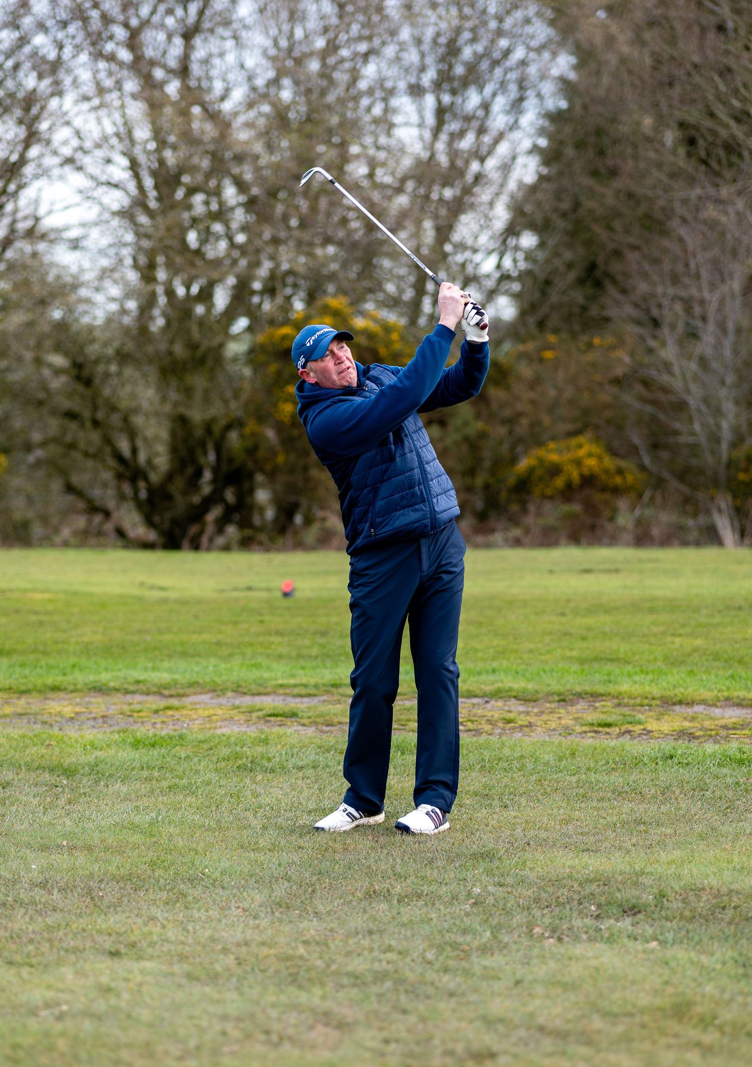 Golfer in blue attire swinging a golf club on a lush green course with trees in the background.