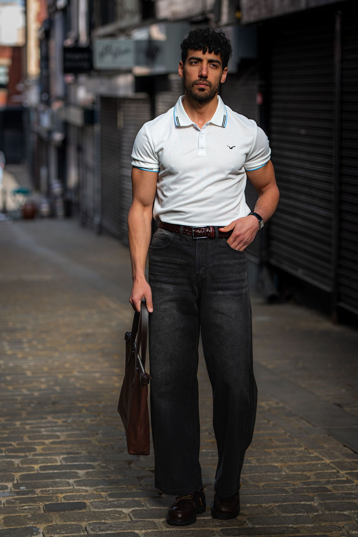 Muscular man in white polo shirt and dark wide-leg jeans holding a brown bag on a cobblestone alley.