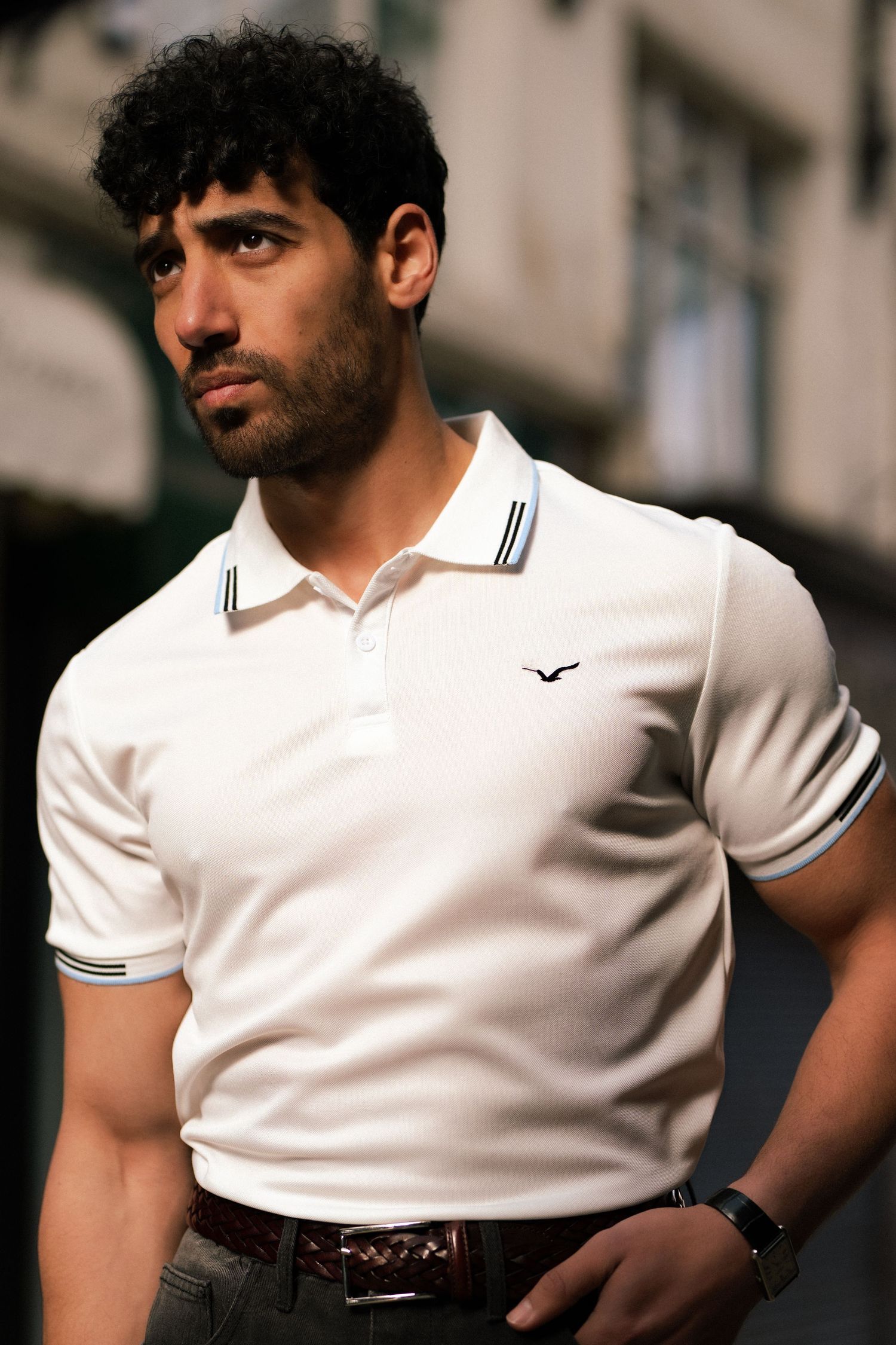 Young man with curly hair wearing a white polo shirt with navy trim, posing outdoors in urban setting.