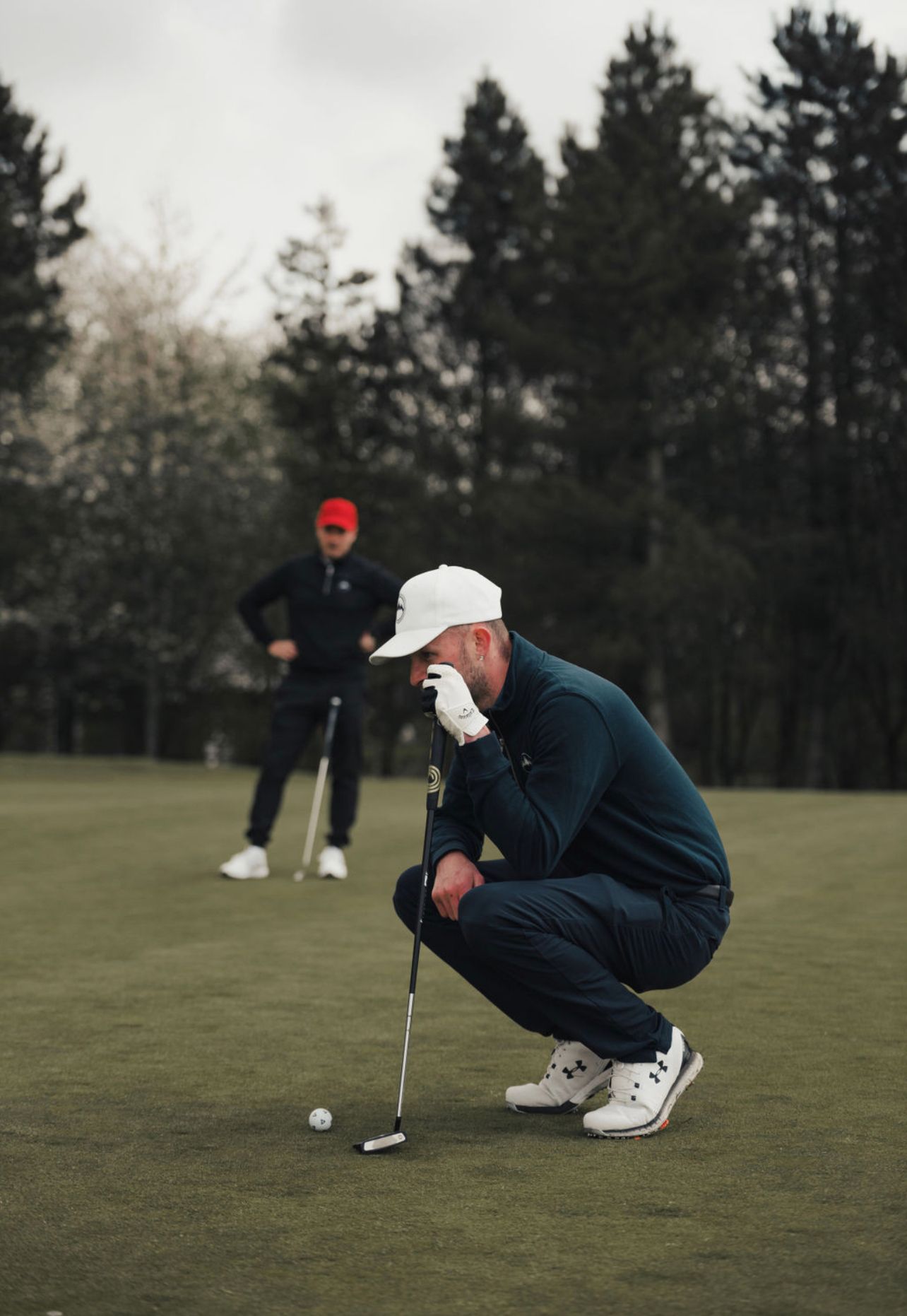 Golfer in dark clothing crouches down to analyze putting line on green golf course.