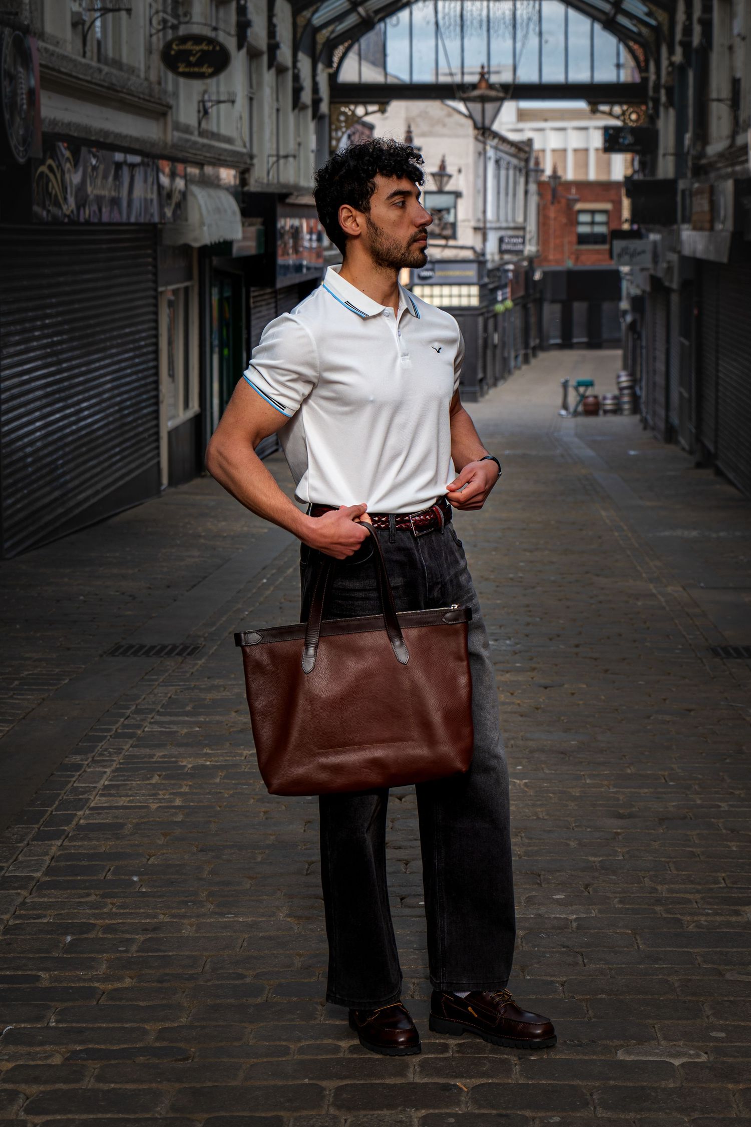 Man in white polo shirt holding brown leather tote bag standing in cobblestone market arcade.