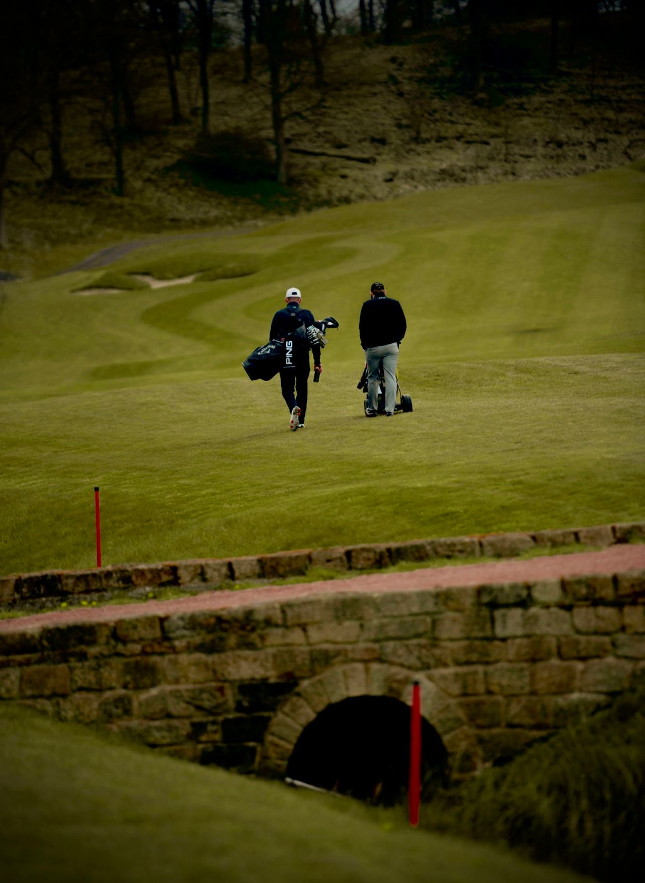 Golfers with bags walk across a stone bridge on a misty golf course with lush green fairways.