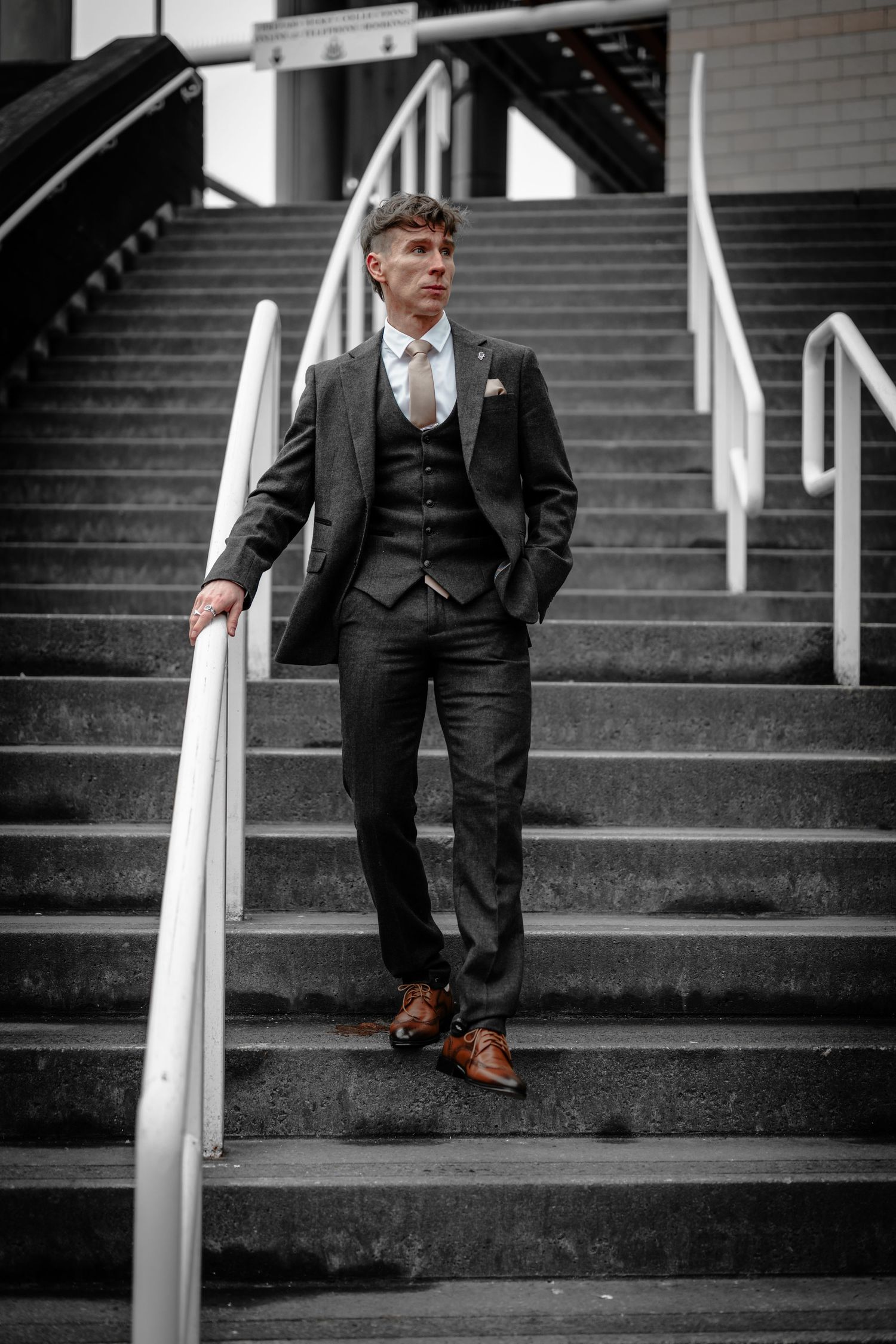 A well-dressed person in a dark three-piece suit and brown leather shoes poses on concrete stairs with white railings.