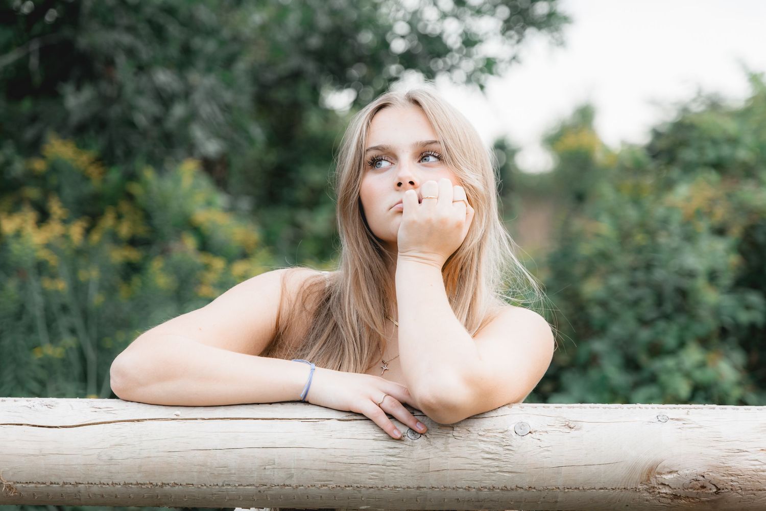 Blonde woman leaning on wooden fence gazing upward in lush green garden setting.