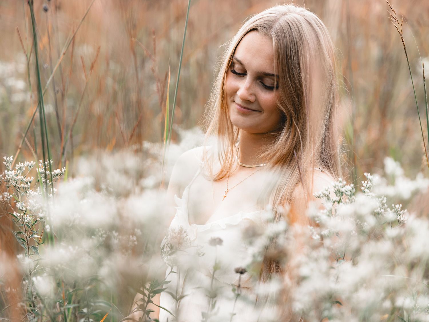 Blonde woman sitting peacefully among white wildflowers in a soft, dreamy outdoor setting.