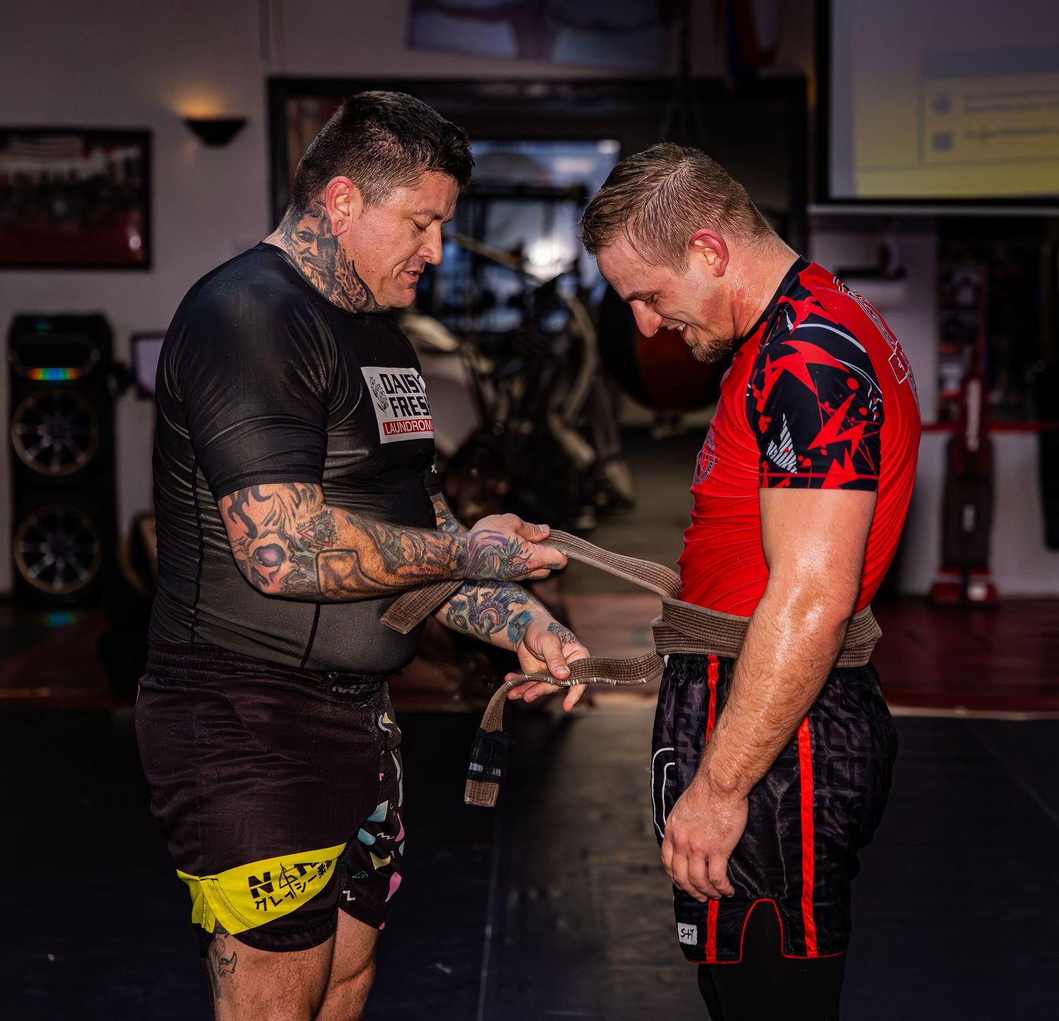 Two MMA fighters wrap hands before a grappling training session in a martial arts gym.