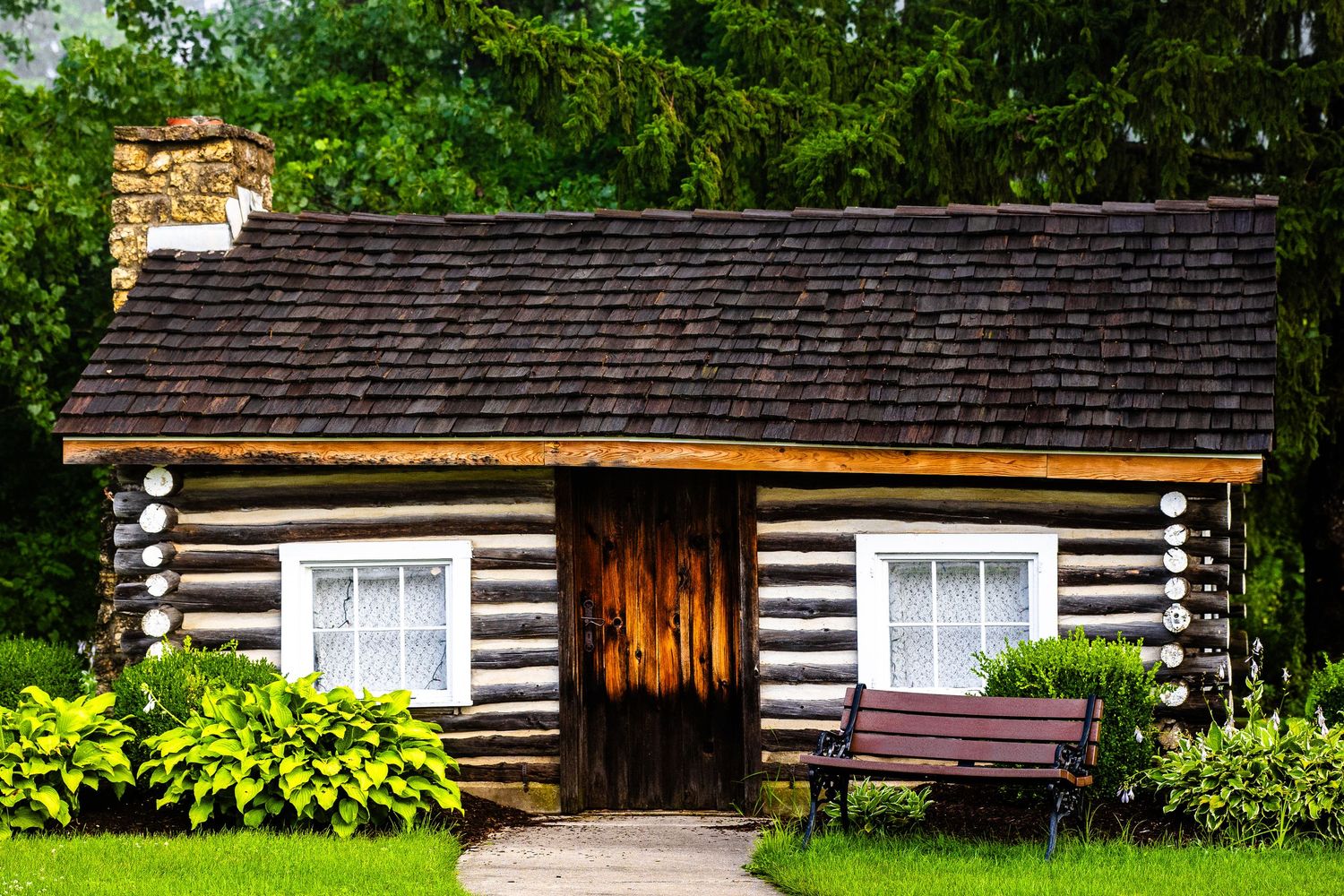 Rustic log cabin with dark wood exterior, white windows, and bench on green lawn.