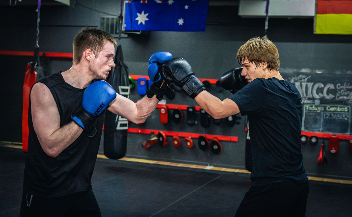 Two male boxers sparring with blue and black gloves in a gym with an Australian flag on the wall.