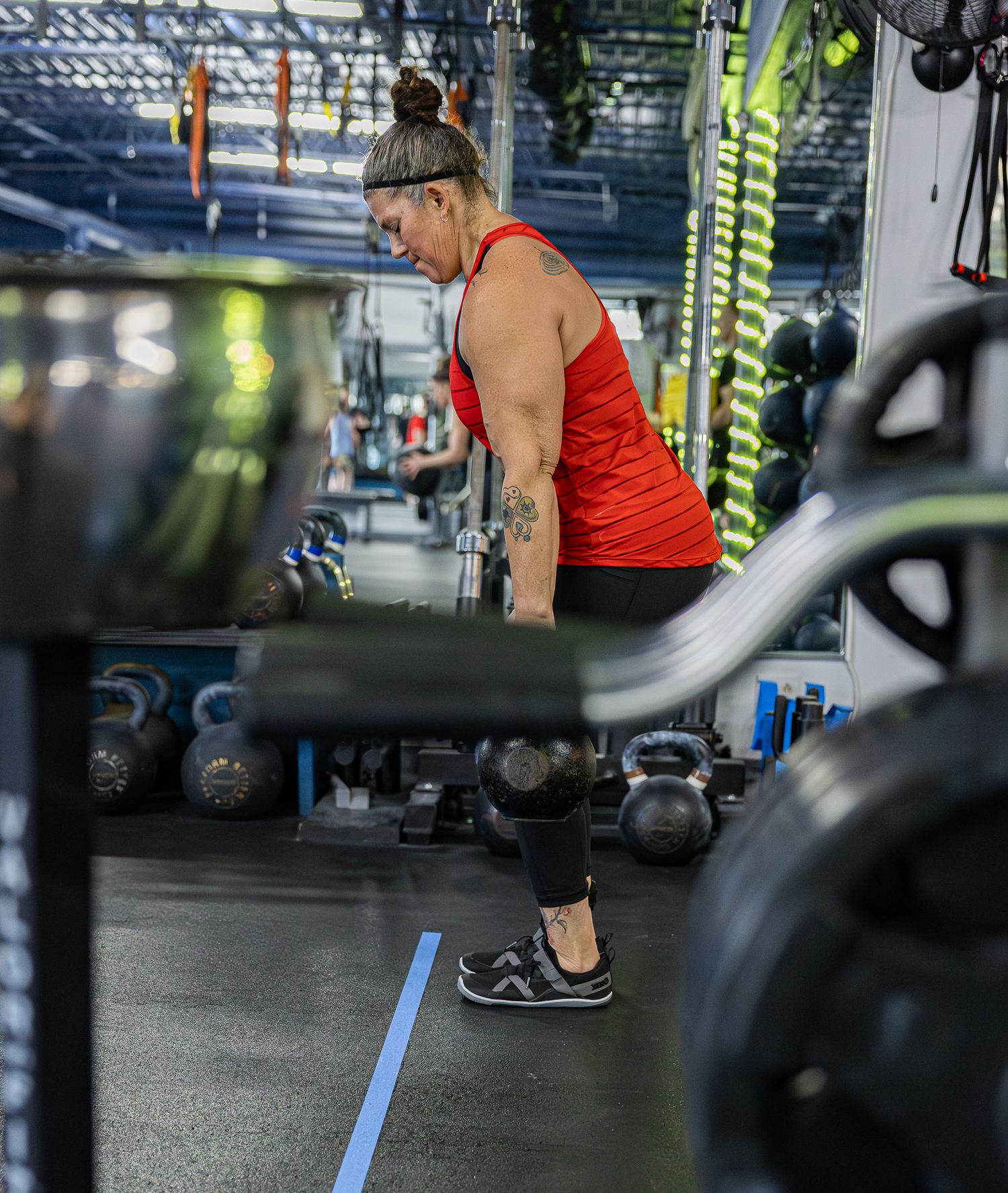 A tattooed woman in a red tank top lifts weights at a well-equipped gym.