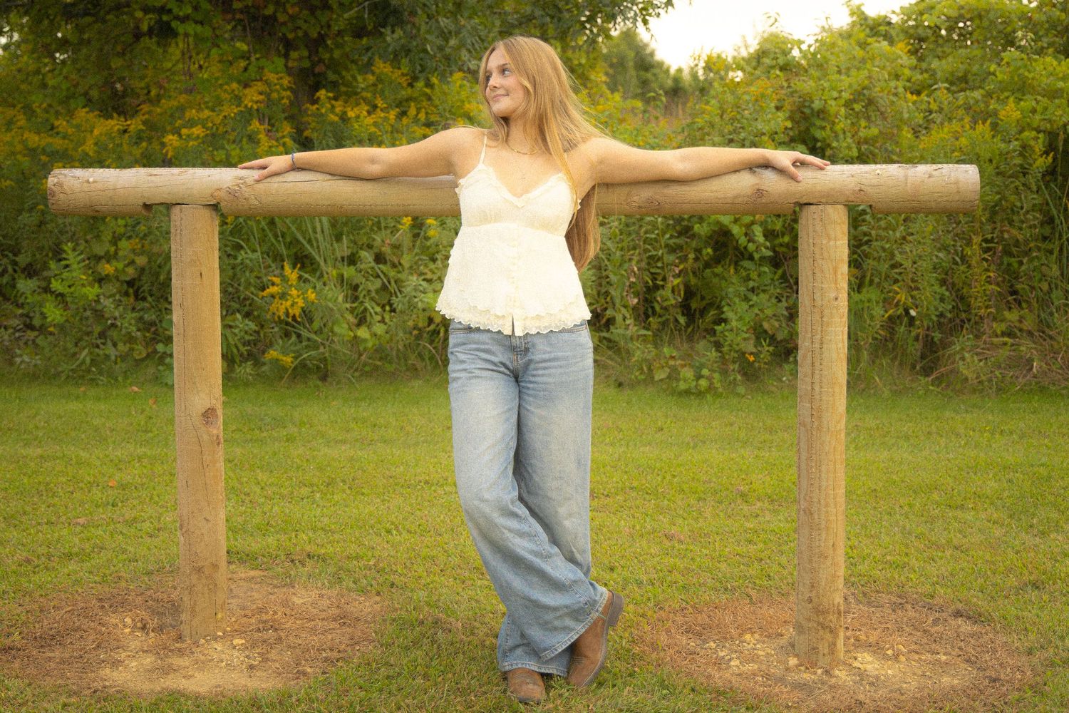 Smiling blonde woman in jeans and white top poses against a rustic wooden fence outdoors.