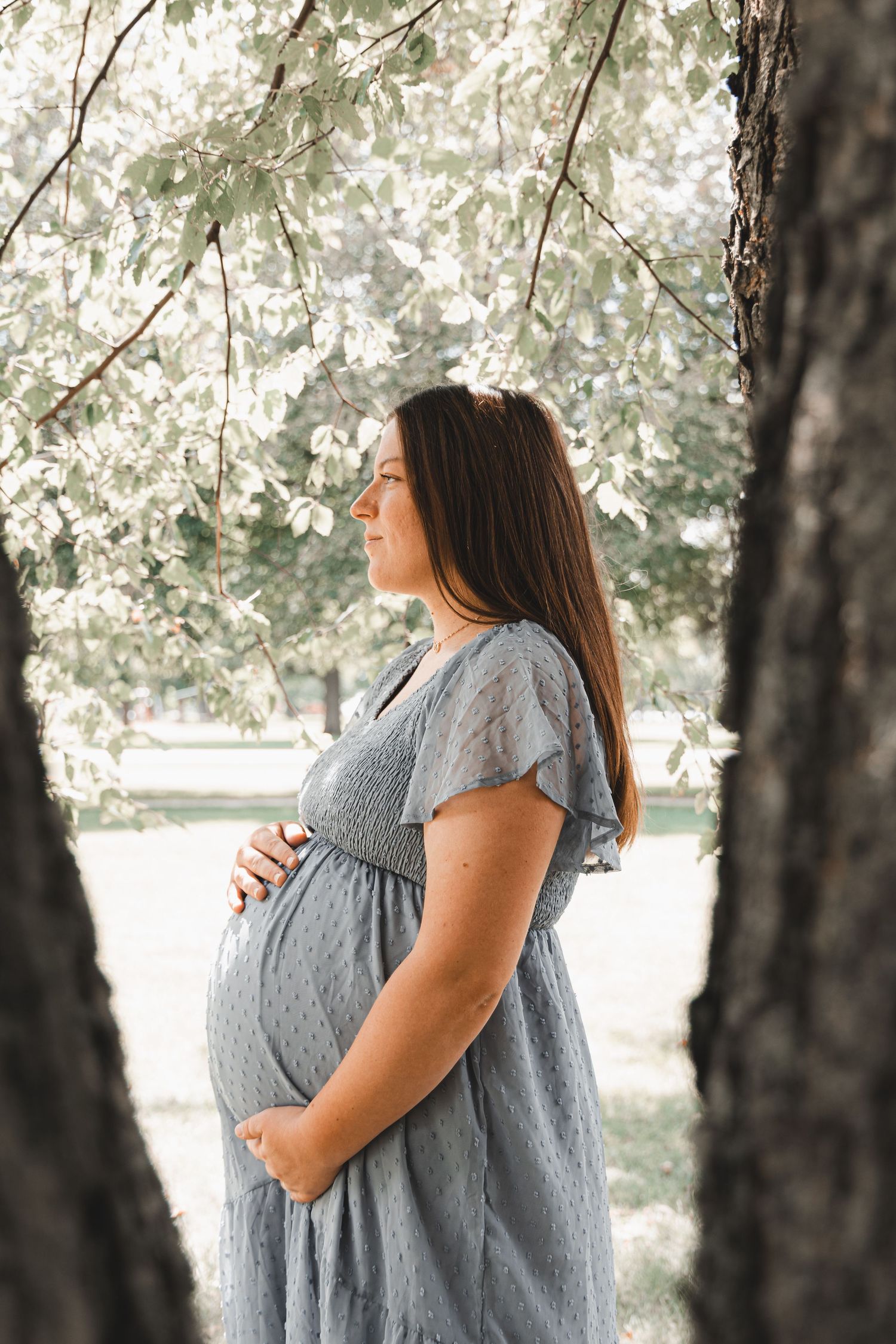 Pregnant woman in a grey dress standing between trees in a blooming park.