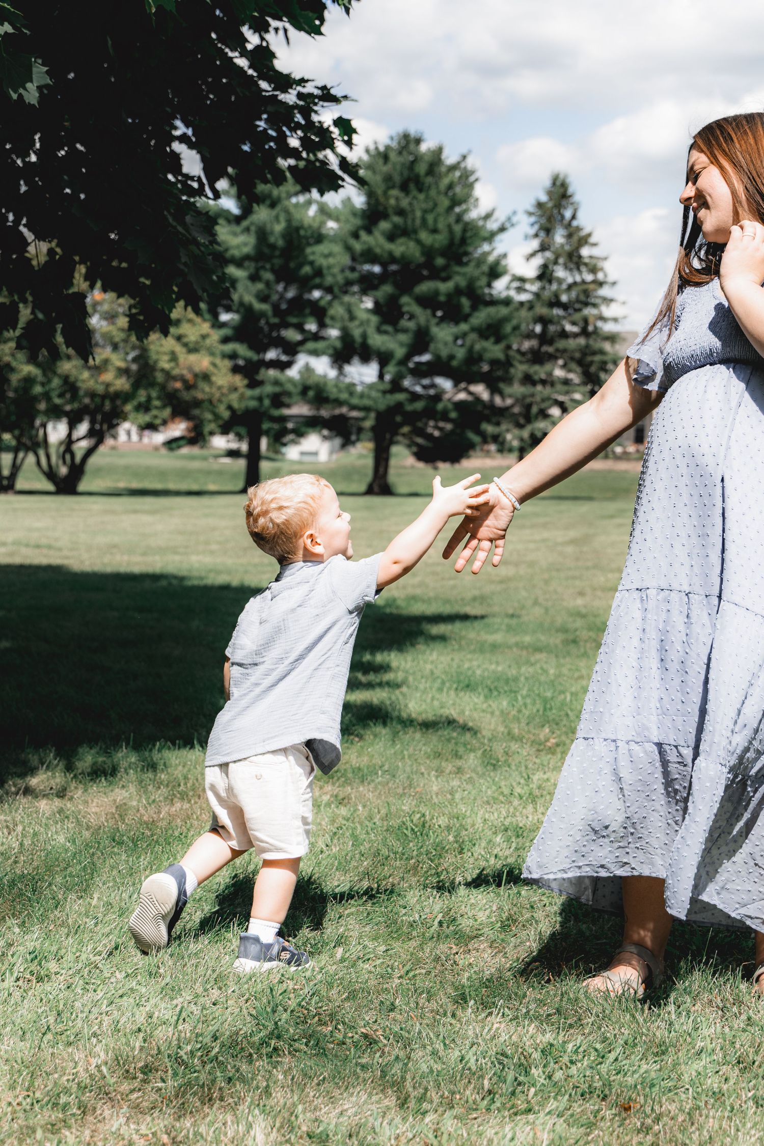 A young blonde toddler holds hands with his pregnant mother while walking together on green grass in a park.
