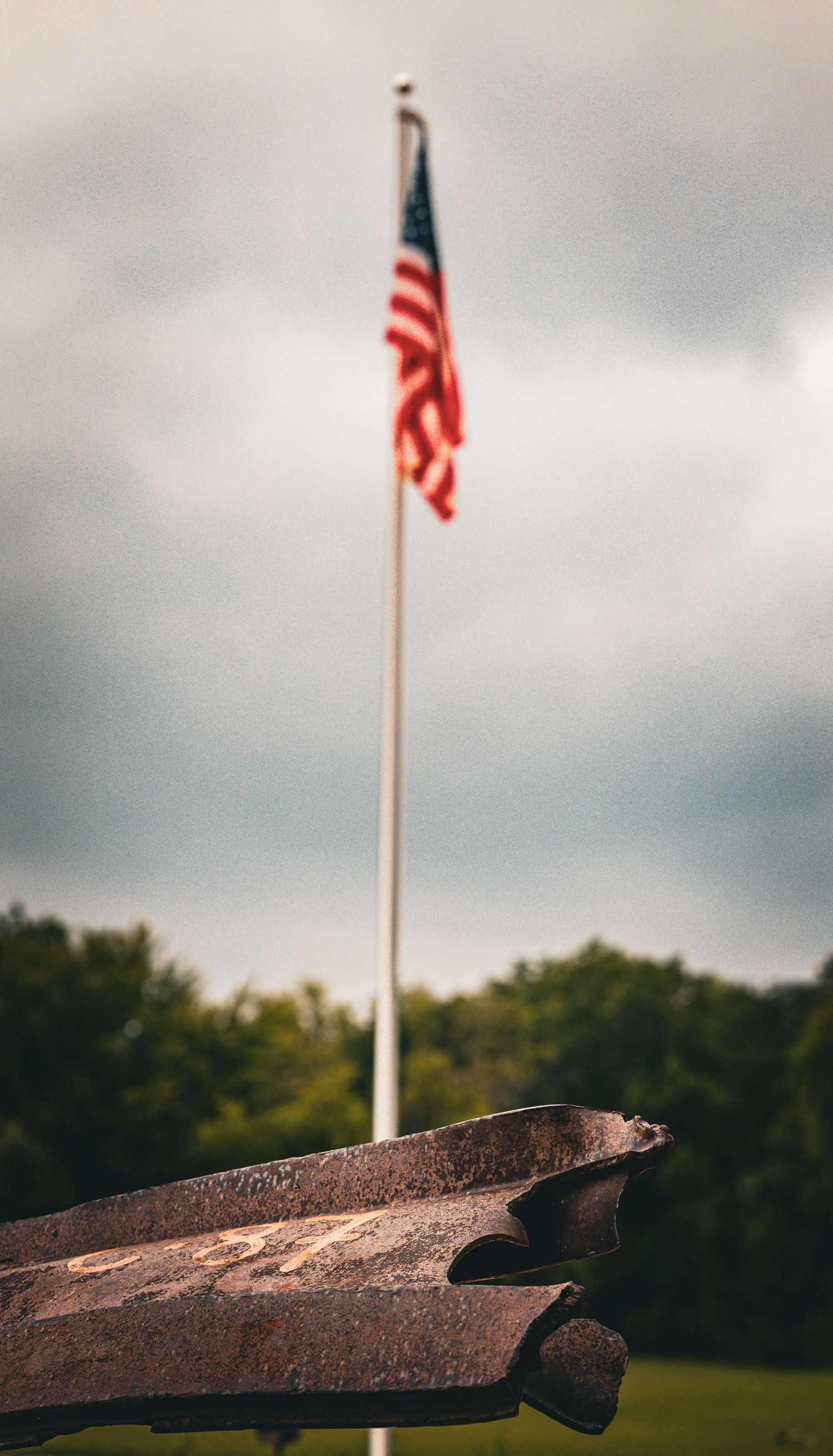 American flag flying on a tall pole behind a rusty metal anchor on a cloudy day.
